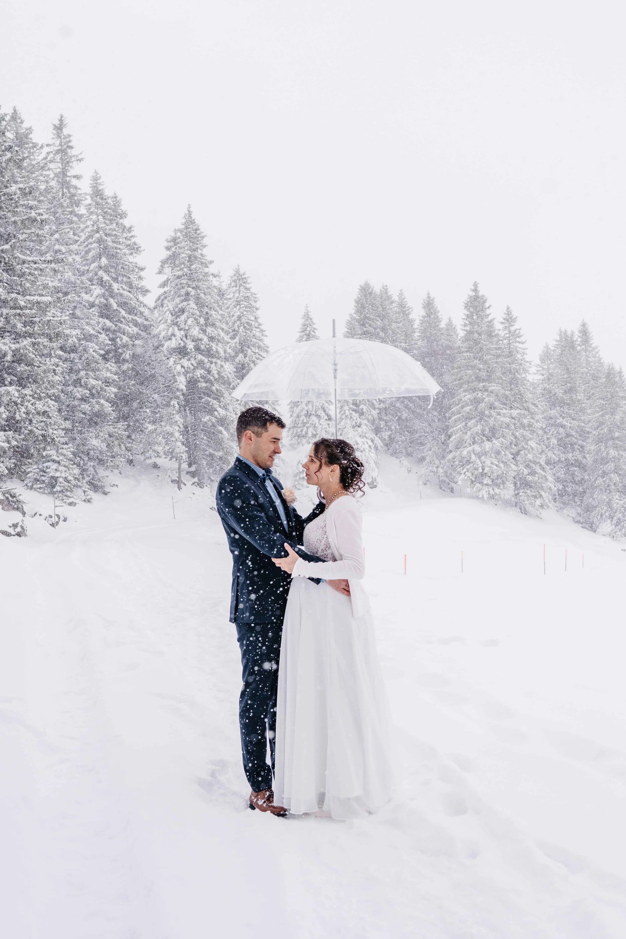 Ein Brautpaar steht in einem verschneiten Winterlandschaft, hält sich unter einem transparenten Regenschirm und lächelt sich an, während Schneeflocken fallen.
