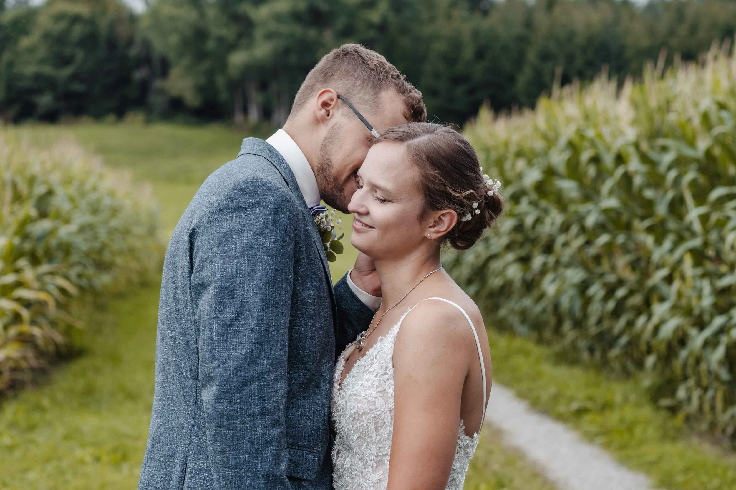Brautpaar umarmt sich in einem Feld, das Bild zeigt den Moment der Liebe. Die Frau trägt ein weisses Hochzeitskleid, der Mann einen blauen Anzug.