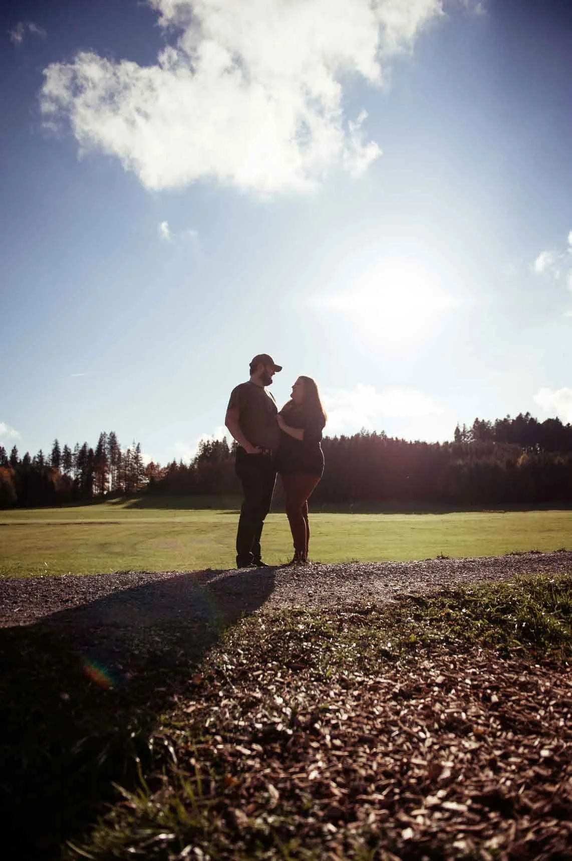 Silhouette eines Paares, das auf einer Wiese im Freien steht, mit der Sonne im Hintergrund und einem blauen Himmel mit wenigen Wolken.