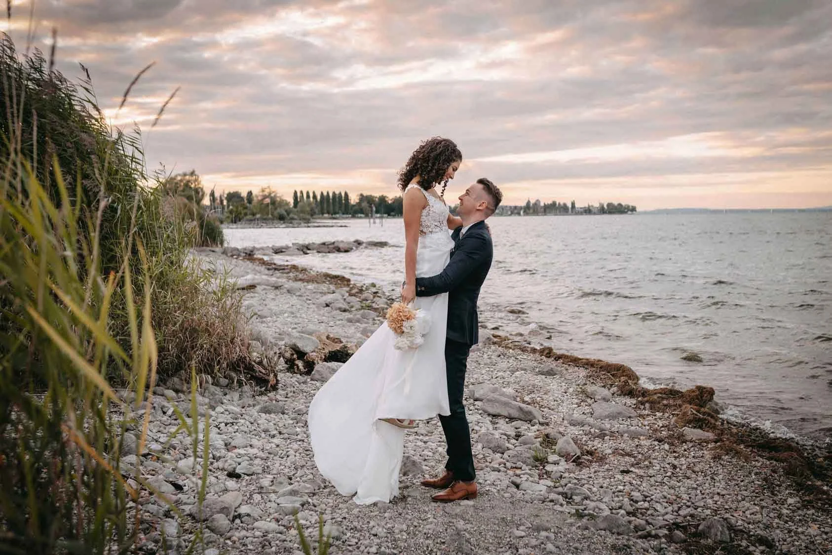 Ein Brautpaar am Strand bei Sonnenuntergang, der Bräutigam hebt die Braut hoch.