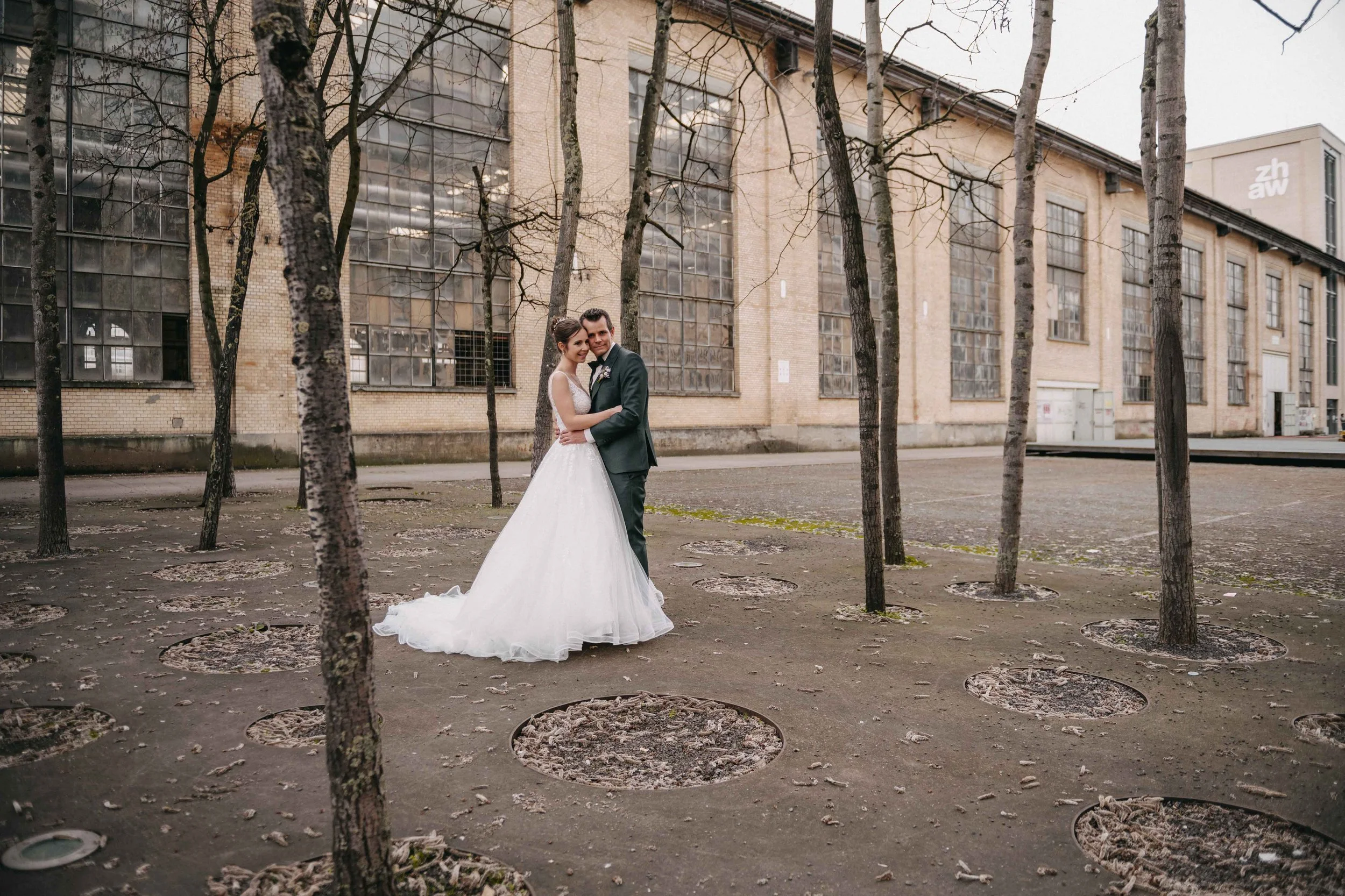 Brautpaar in Hochzeitskleidung, umarmend und lächelnd, im Freien vor einem alten Backsteingebäude mit grossen Fenster.