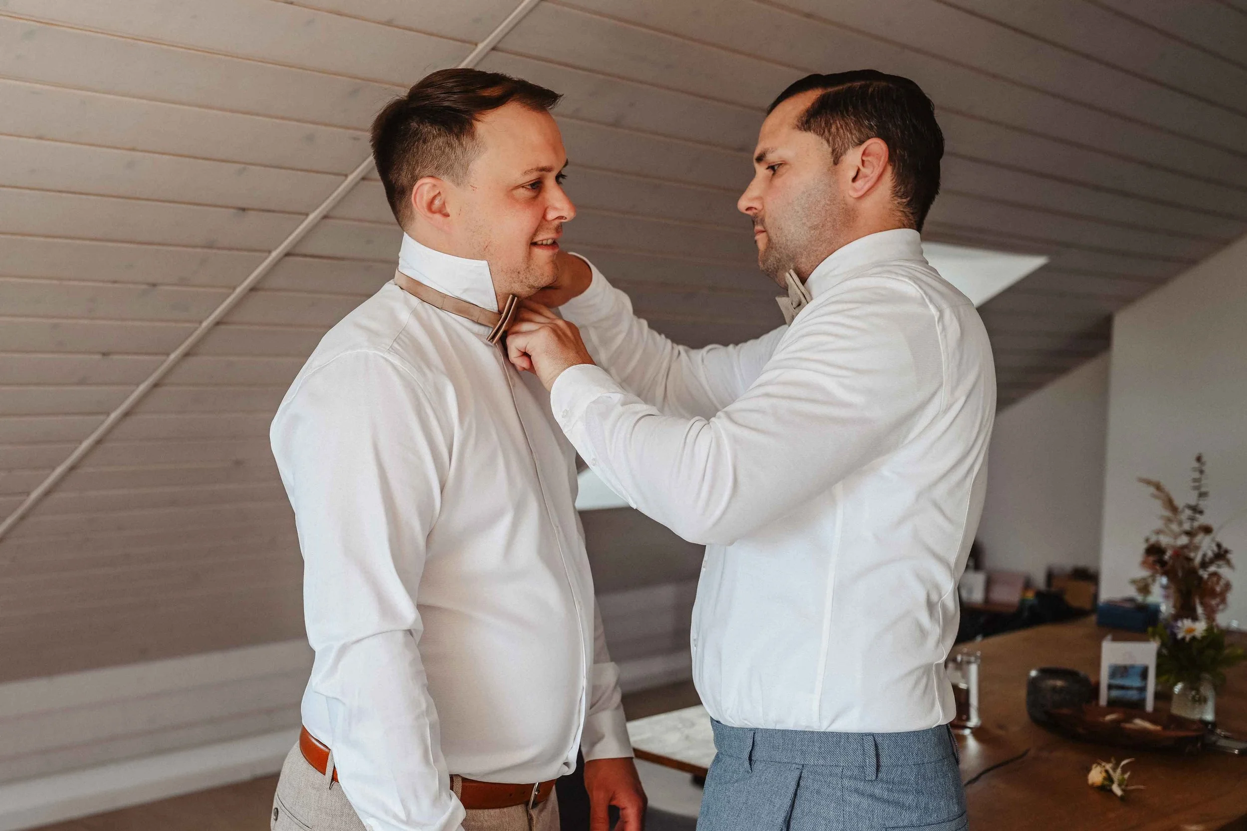 Bräutigam und Trauzeuge stehen sich gegenüber in einem Raum mit Holzdecke. Einer hilft dem anderen, einen Anzug zu richten.