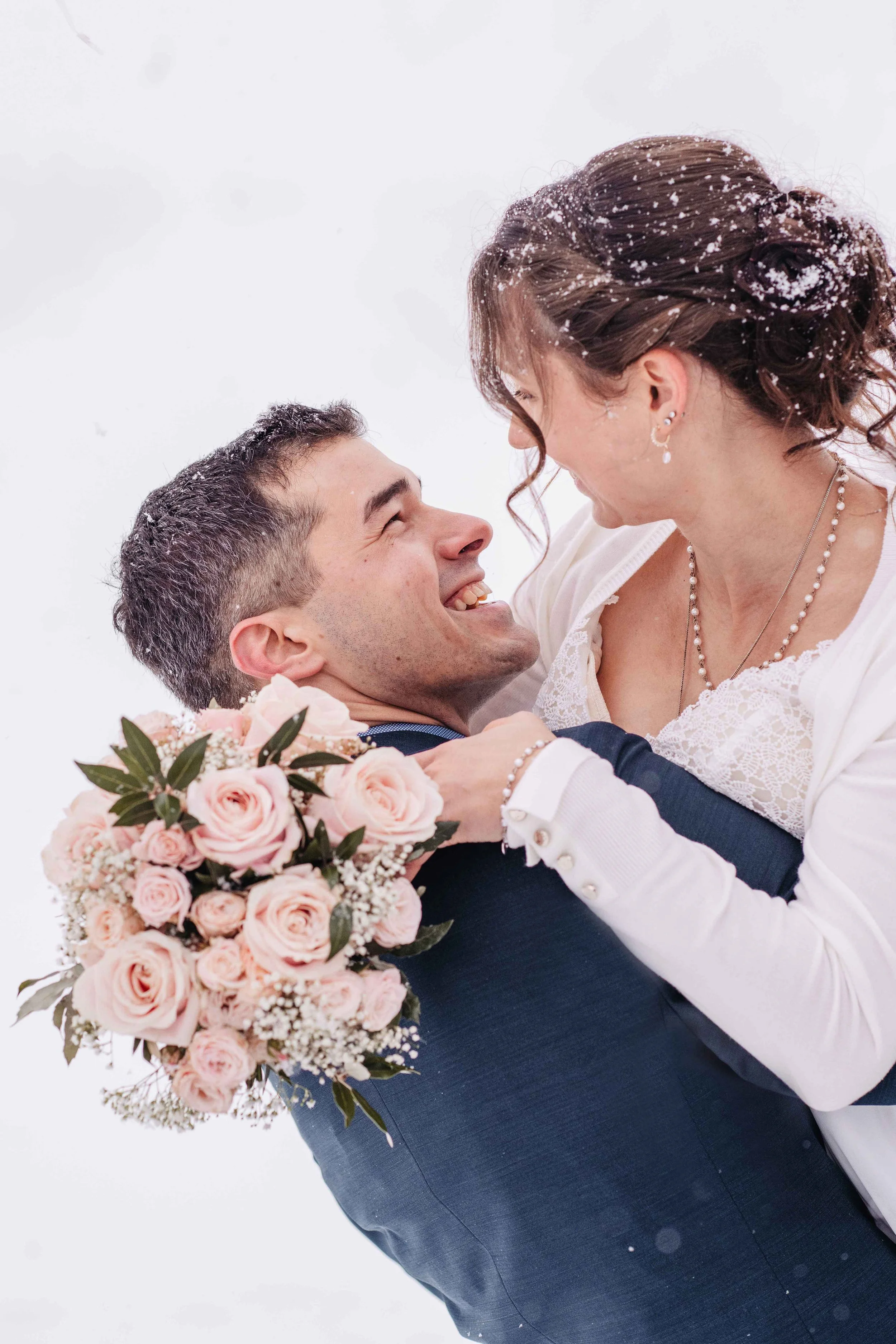 Das Brautpaar, umarmt sich lächelnd in einer verschneiten Winterlandschaft und schauen sich in die Augen.