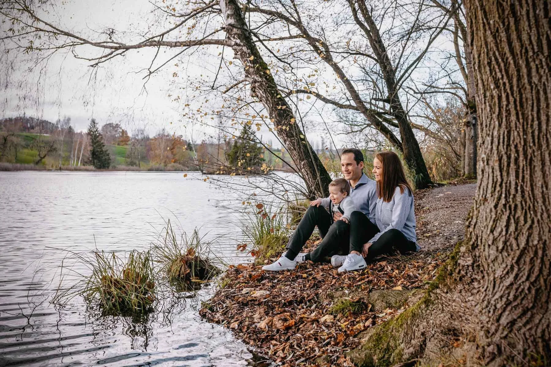 Ein glückliche Familie sitzt am Ufer eines Weihers. Umgeben von herbstlicher Landschaft und Bäumen, geniessen sie die Natur. Die Eltern halten den kleinen Jungen, während alle lächeln.