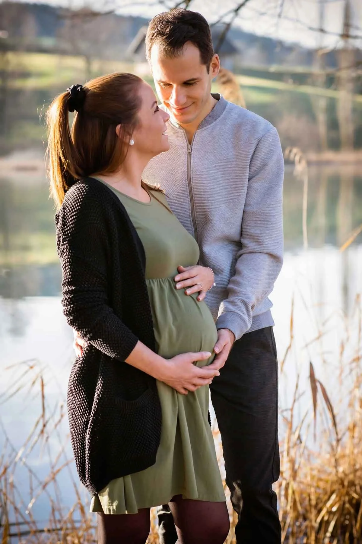 Ein schwangertes Paar steht an einem See in der Natur, sie hält ihren Babybauch und er legt seine Hand darauf. Sie schauen sich liebevoll an.