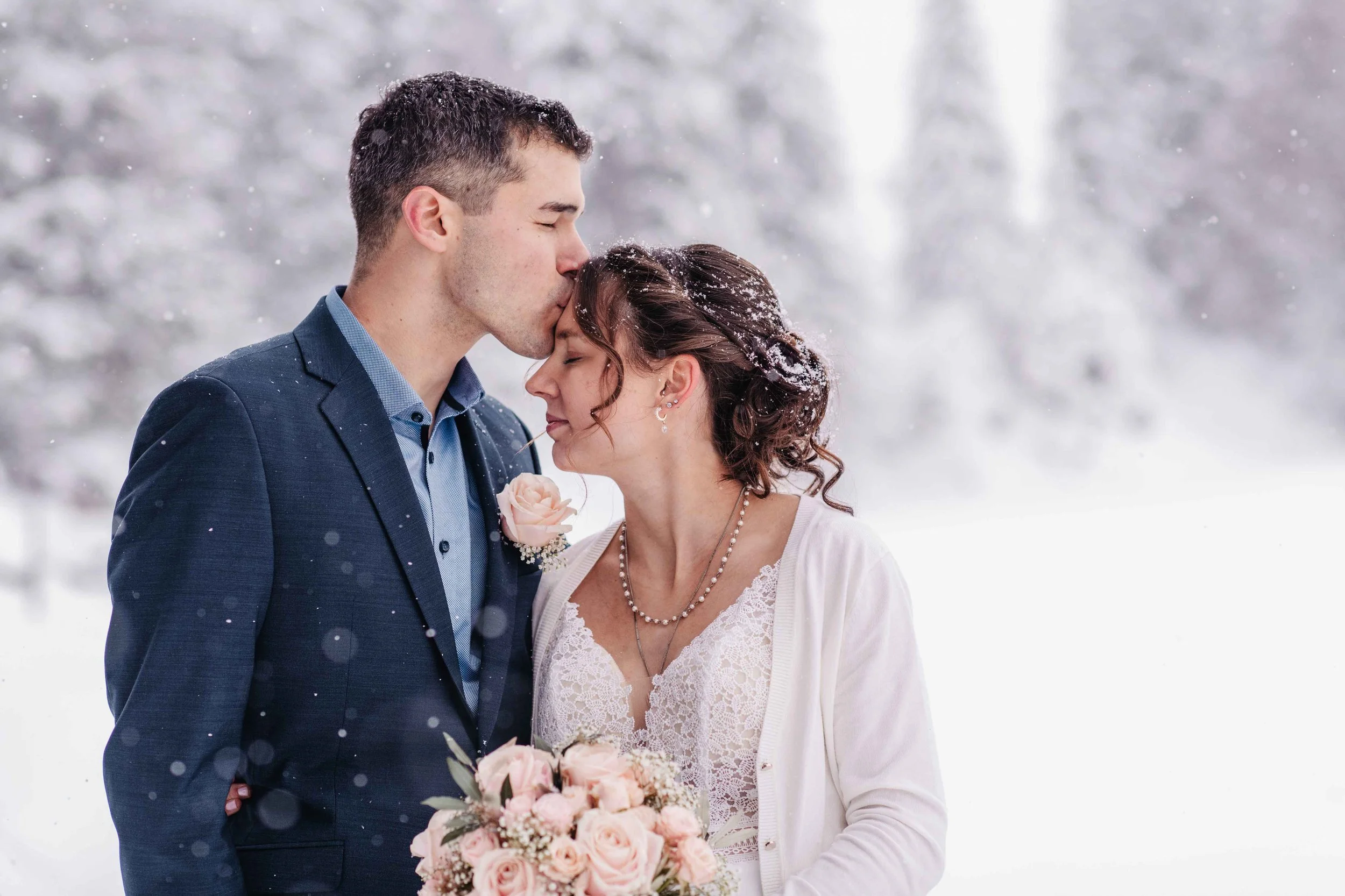 Ein Paar steht im Schnee, der Mann küsst die Braut auf die Stirn, Braut hält einen Blumenstrauss aus weissen und rosafarbenen Rosen.