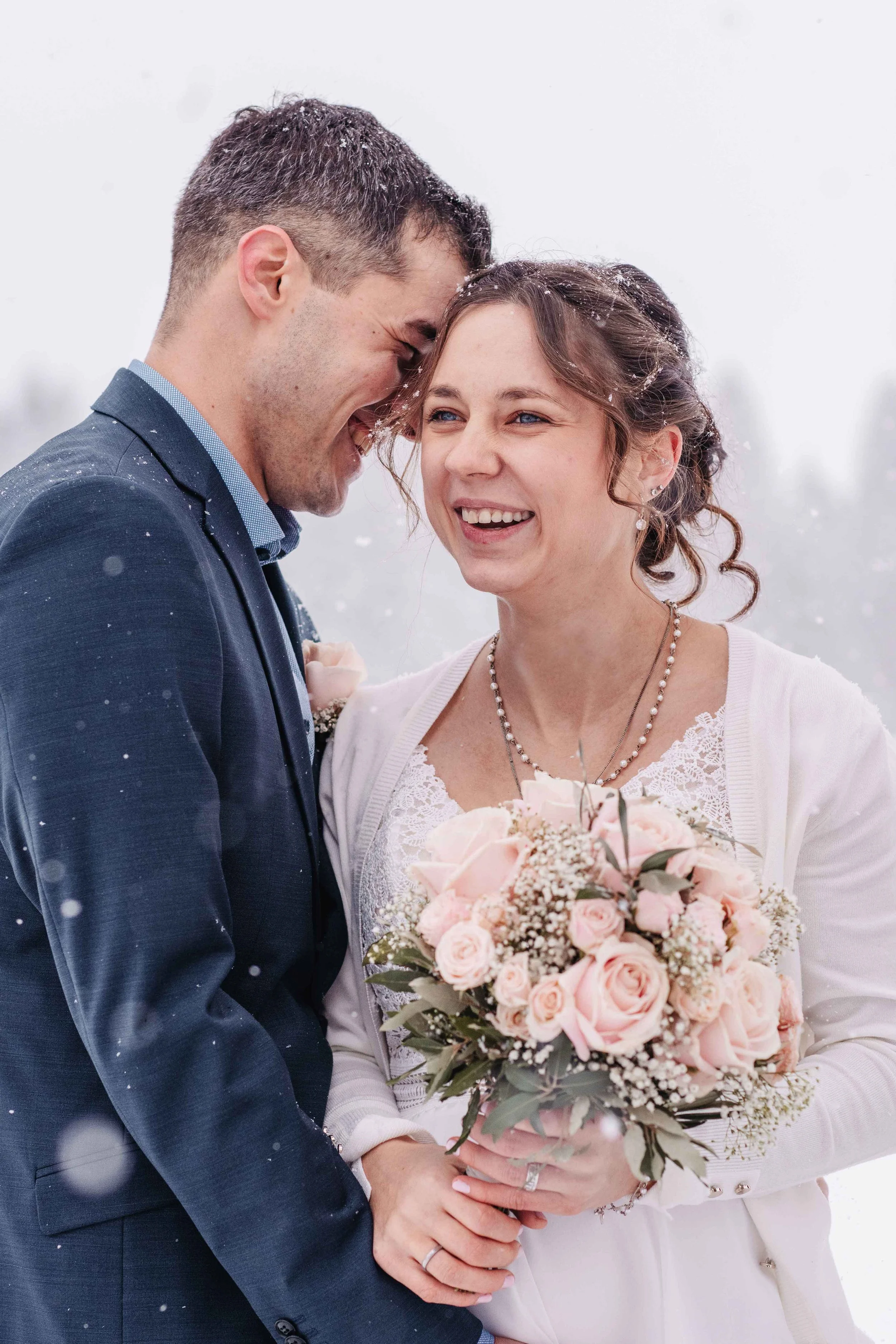 Ein lächelndes Paar bei einer Winterhochzeit, die Frau hält einen Blumenstrauss, beide tragen Hochzeitsringe.