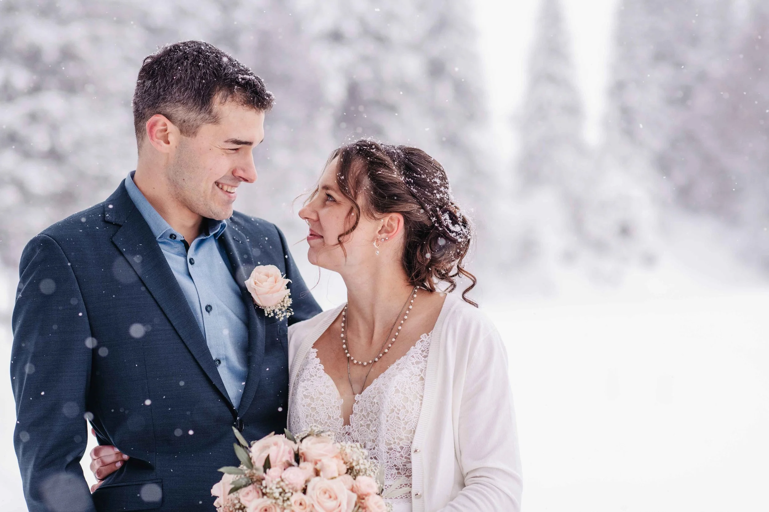 Ein Paar bei einer winterlichen Hochzeitsaufnahme, der Mann trägt einen Anzug und die Frau ein Kleid mit einem Brautstrauss, schneebedeckter Hintergrund.