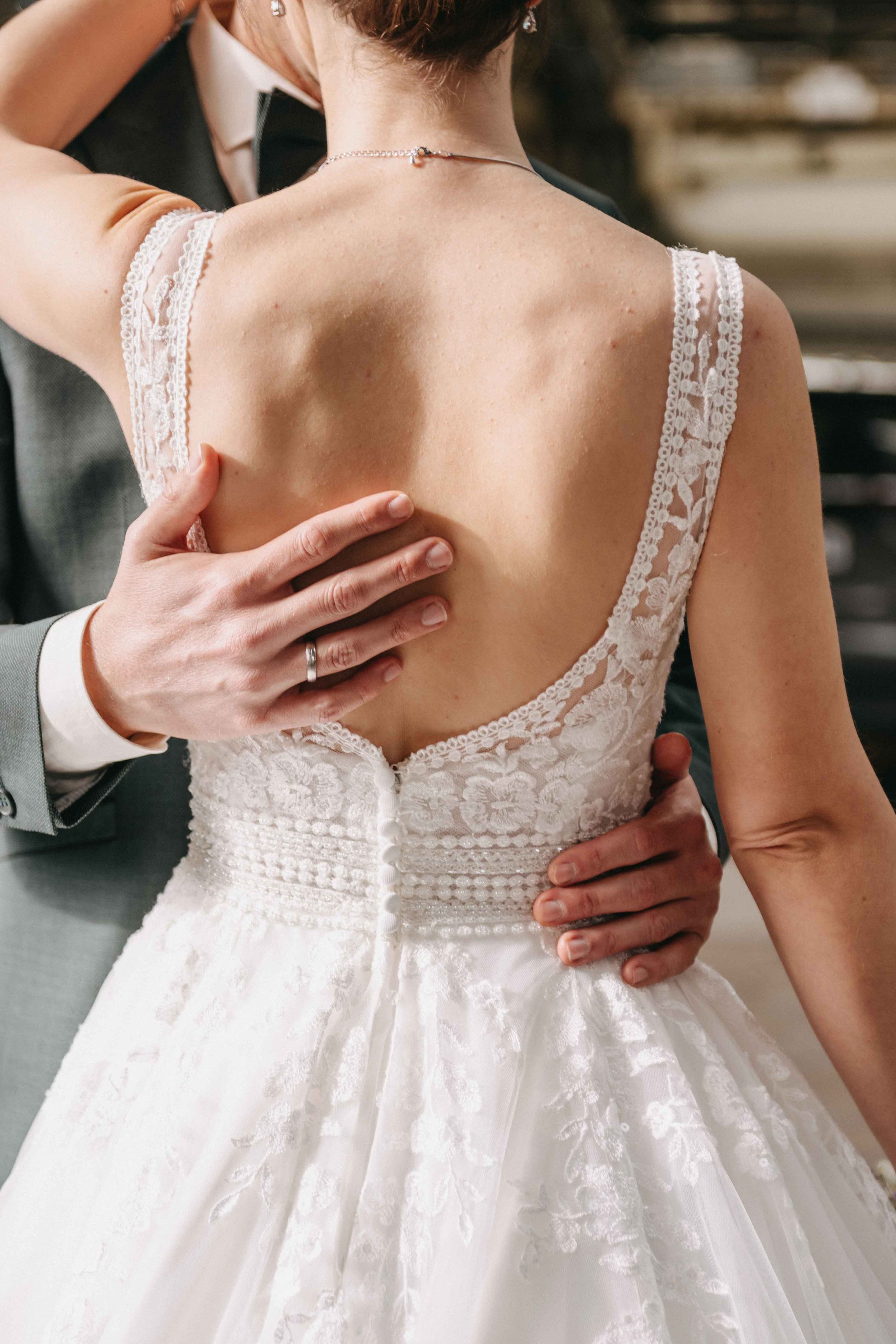 Nahaufnahme eines Brautkleides bei einer Hochzeit. Die Braut trägt ein weisses Kleid mit lila Spitzen und einer tiefen Rückenausschnitt, während der Bräutigam, an sie heranlehnt.