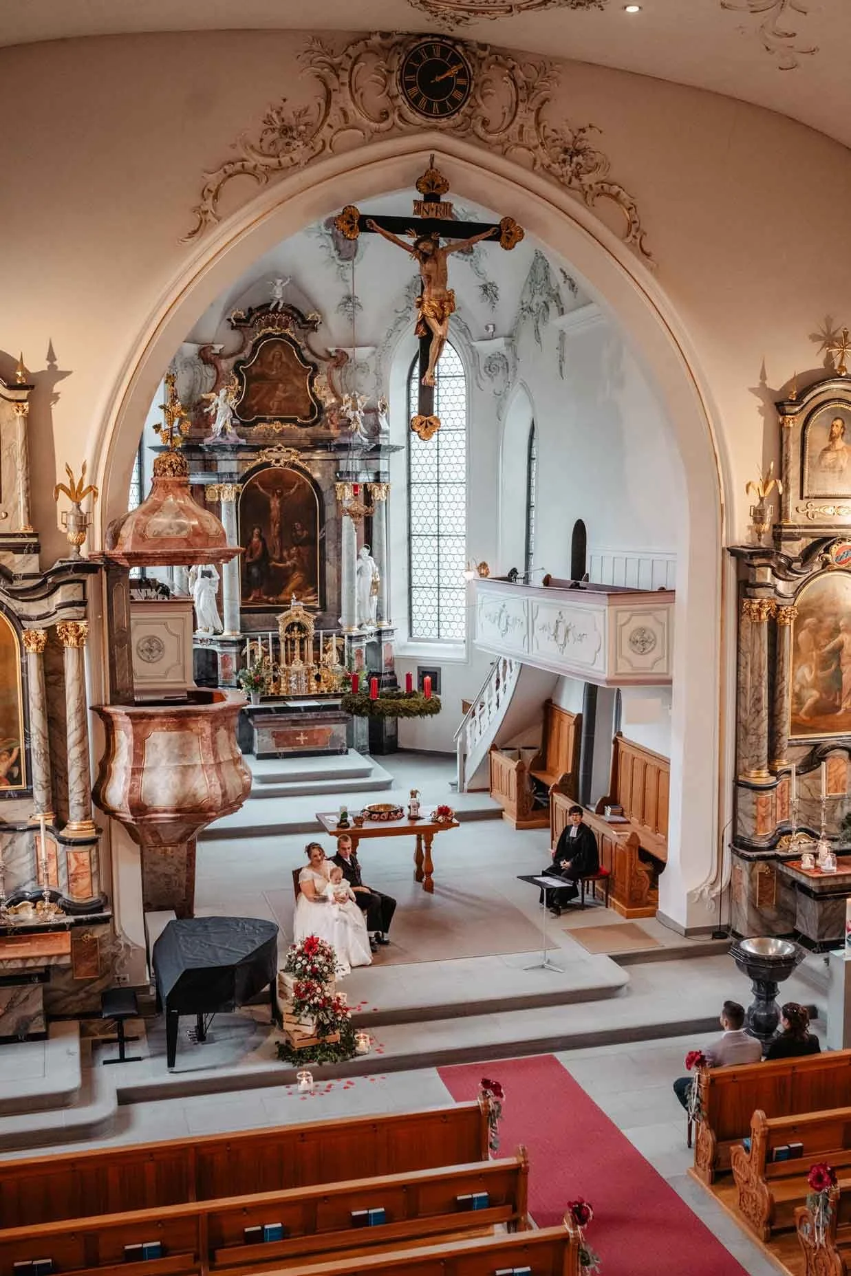 Hochzeit in einer barocken Kirche mit Braut, Bräutigam, einem Kind, einem Pfarrer, und Gästen, alles in festlicher Stimmung bei einer kirchlichen Trauung.