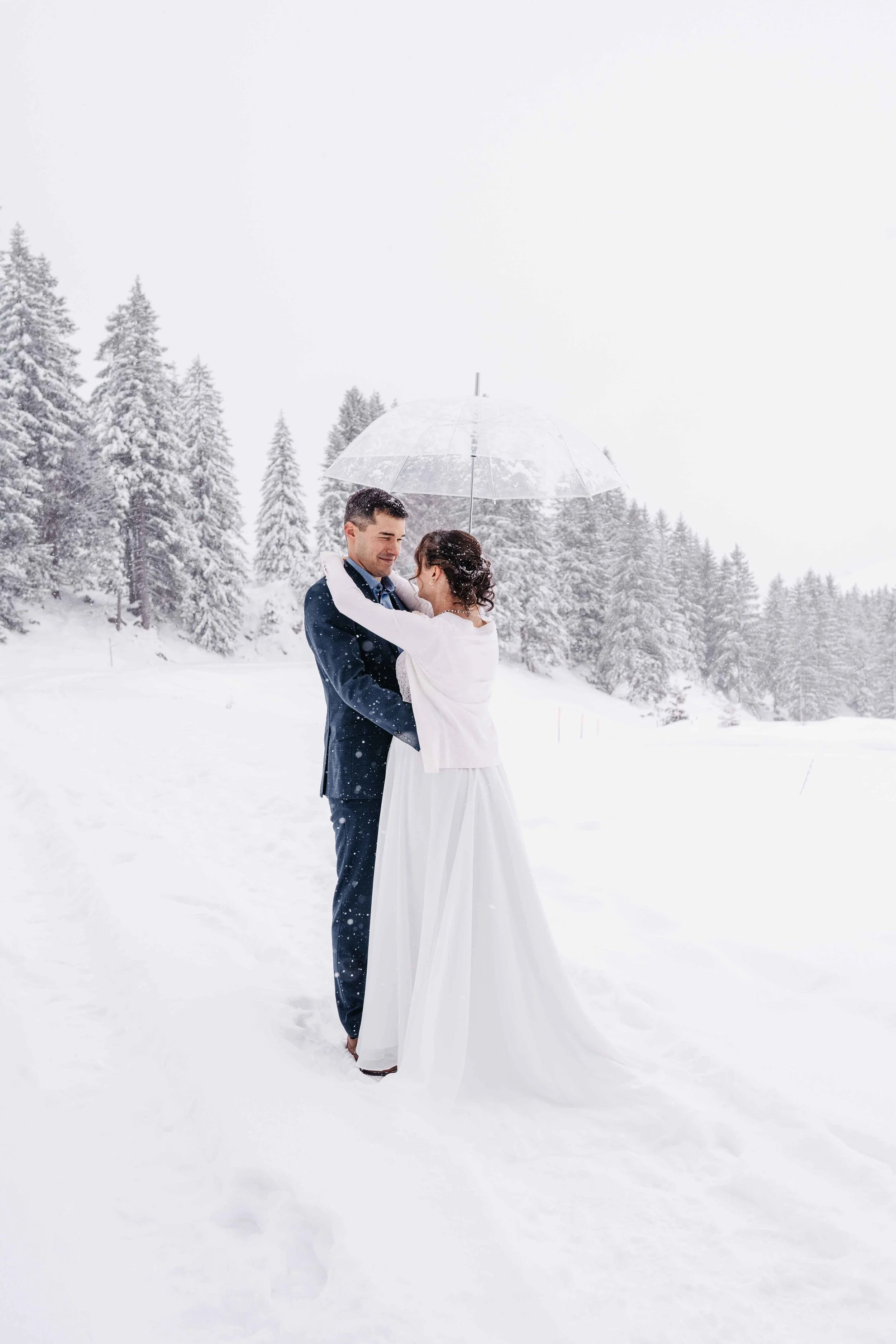 Ein Paar in Hochzeitskleidung unter einem transparenten Regenschirm in einer verschneiten Winterlandschaft, umgeben von schneebedeckten Tannenbäumen.