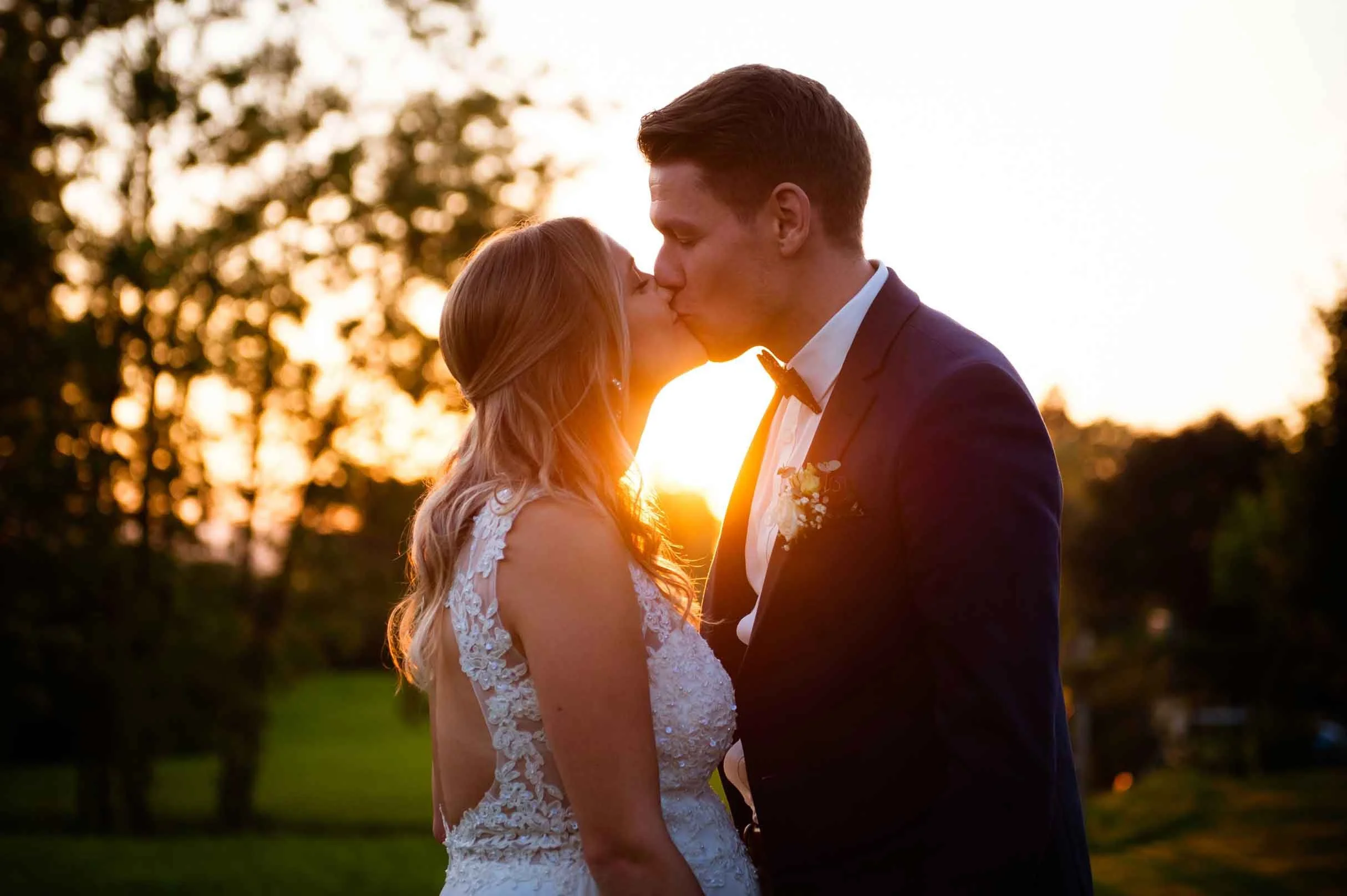 Ein Brautpaar küsst sich bei Sonnenuntergang im Freien, umgeben von Bäumen.