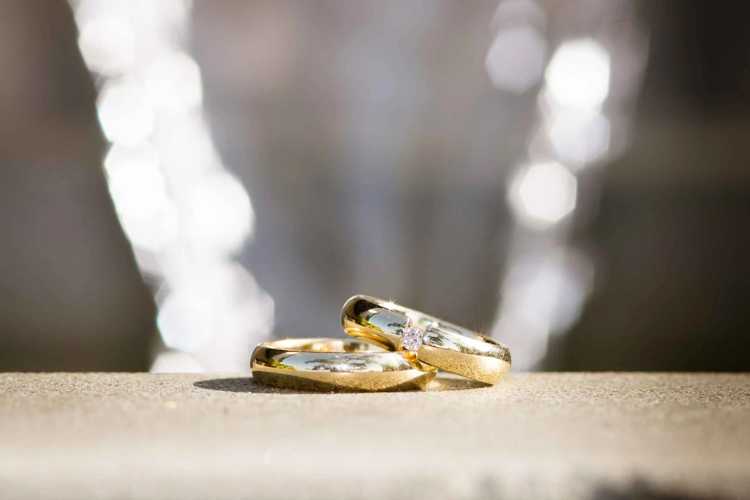 Goldene Eheringe, einer mit einem Diamanten, auf einer Sandoberfläche, unscharfer Hintergrund mit Lichteffekten.