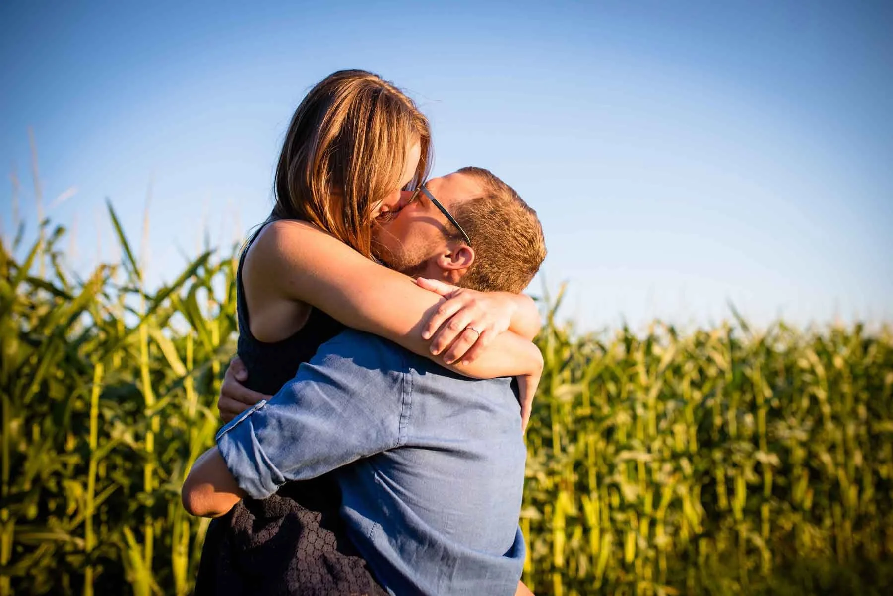 Ein Paar umarmt sich im Freien bei Sonnenuntergang, im Hintergrund Maisfelder, blauer Himmel.