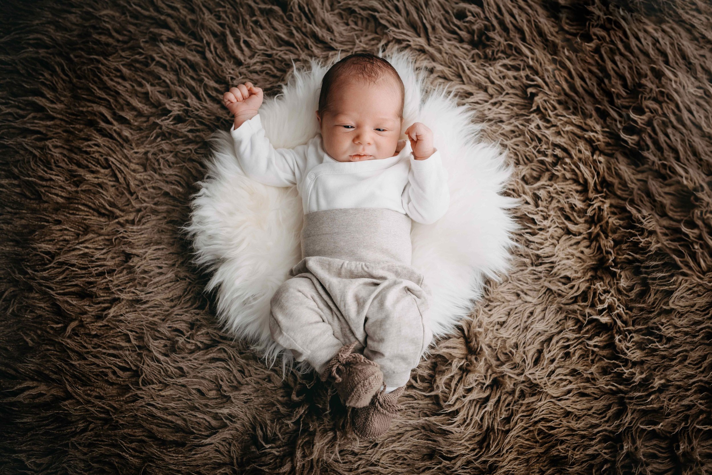 Newborn Baby in weissem Oberteil und grauen Hosen, liegt auf einem flauschigen braunen Teppich.