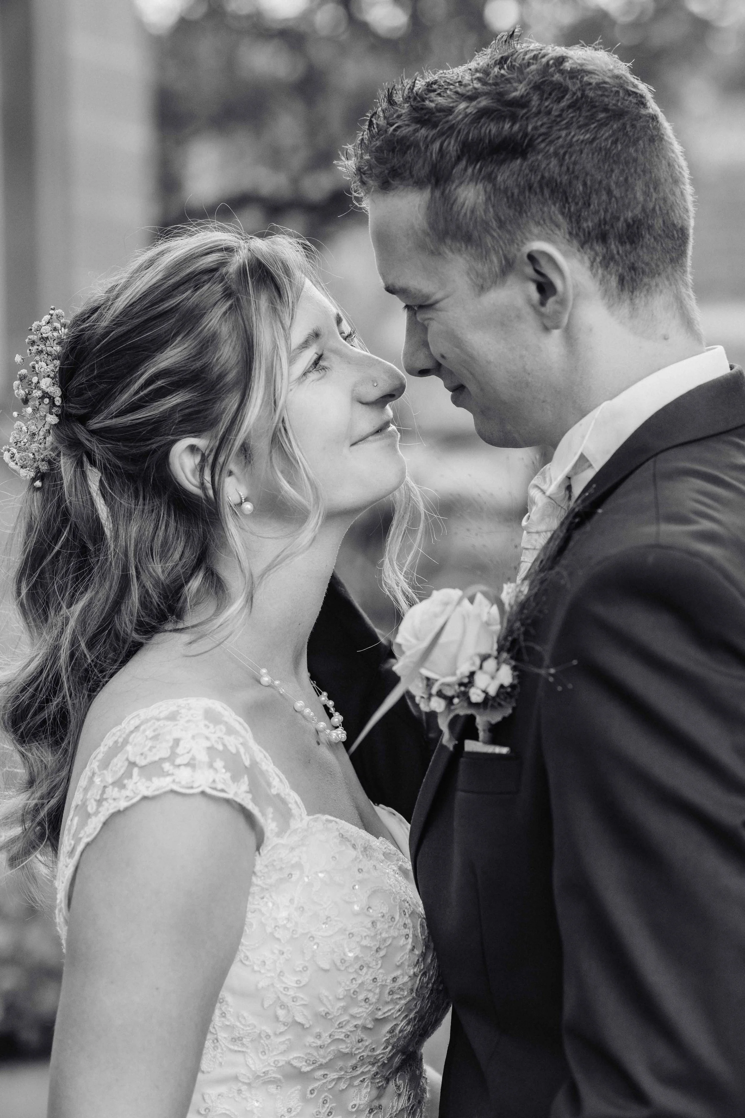 Brautpaar steht sich bei einer Hochzeit gegenüber, lächelt und blickt sich tief in die Augen. Die Braut trägt ein Spitzenkleid mit Perlen, Haarschmuck und Perlenkette, der Bräutigam trägt einen schwarzen Anzug mit einer Rose im Revers.