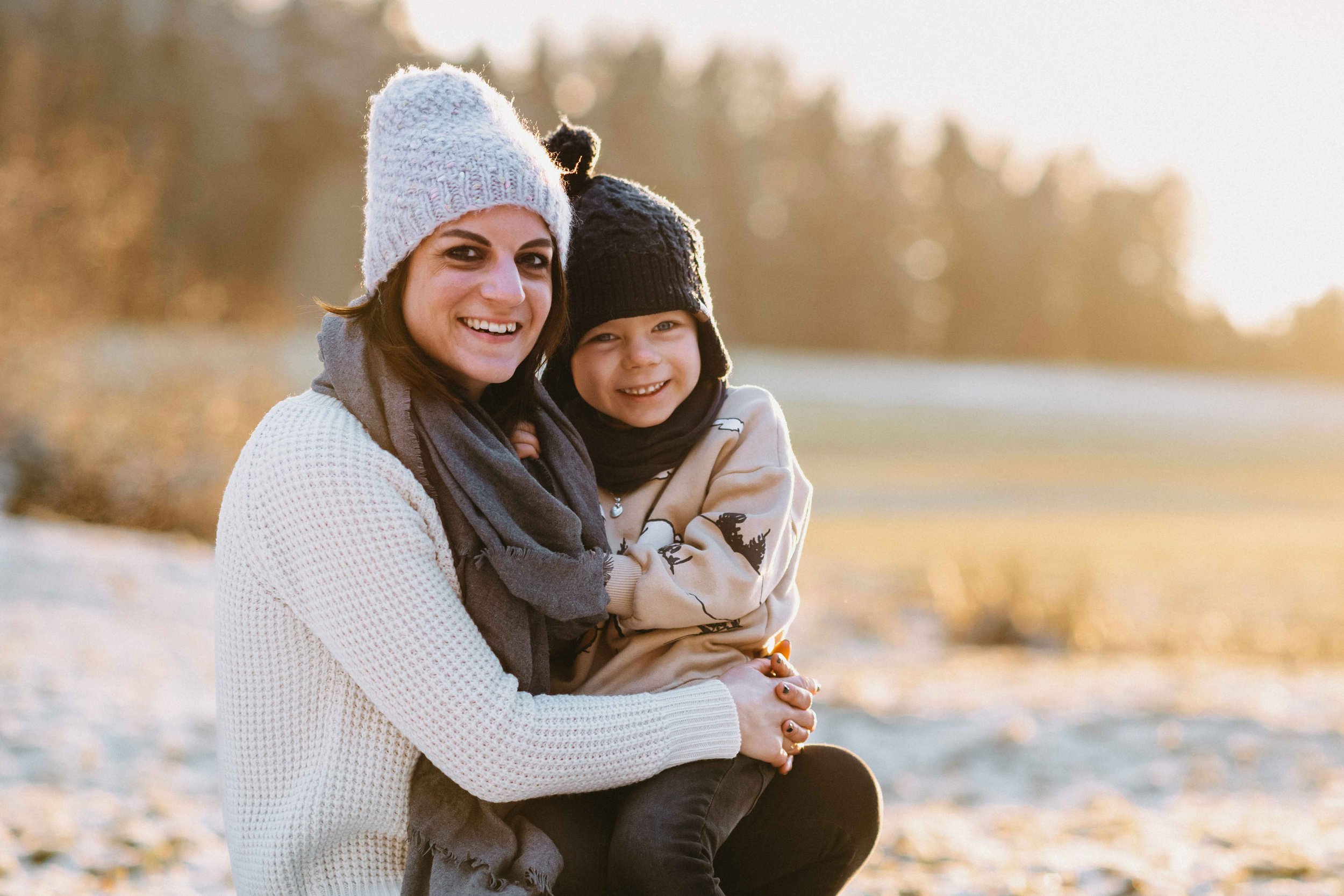 Eine Mama und ihr Kind in Winterkleidung lächeln und umarmen sich im Freien. Sie tragen Mützen und Schals.