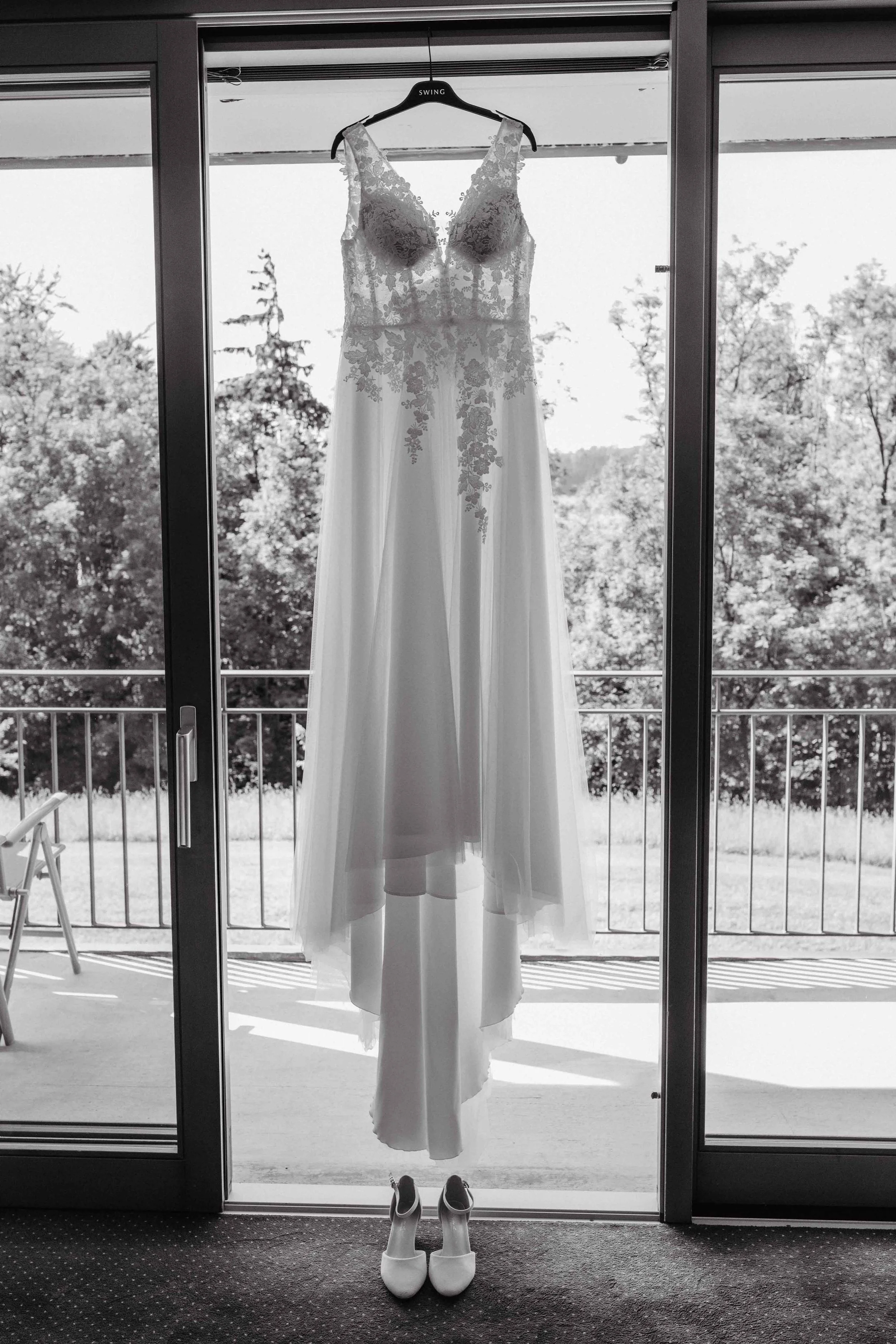 Brautkleid mit weissen Heels, aufgehängt an einem Balkon vor einer Fensterfront mit Blick auf Bäume.