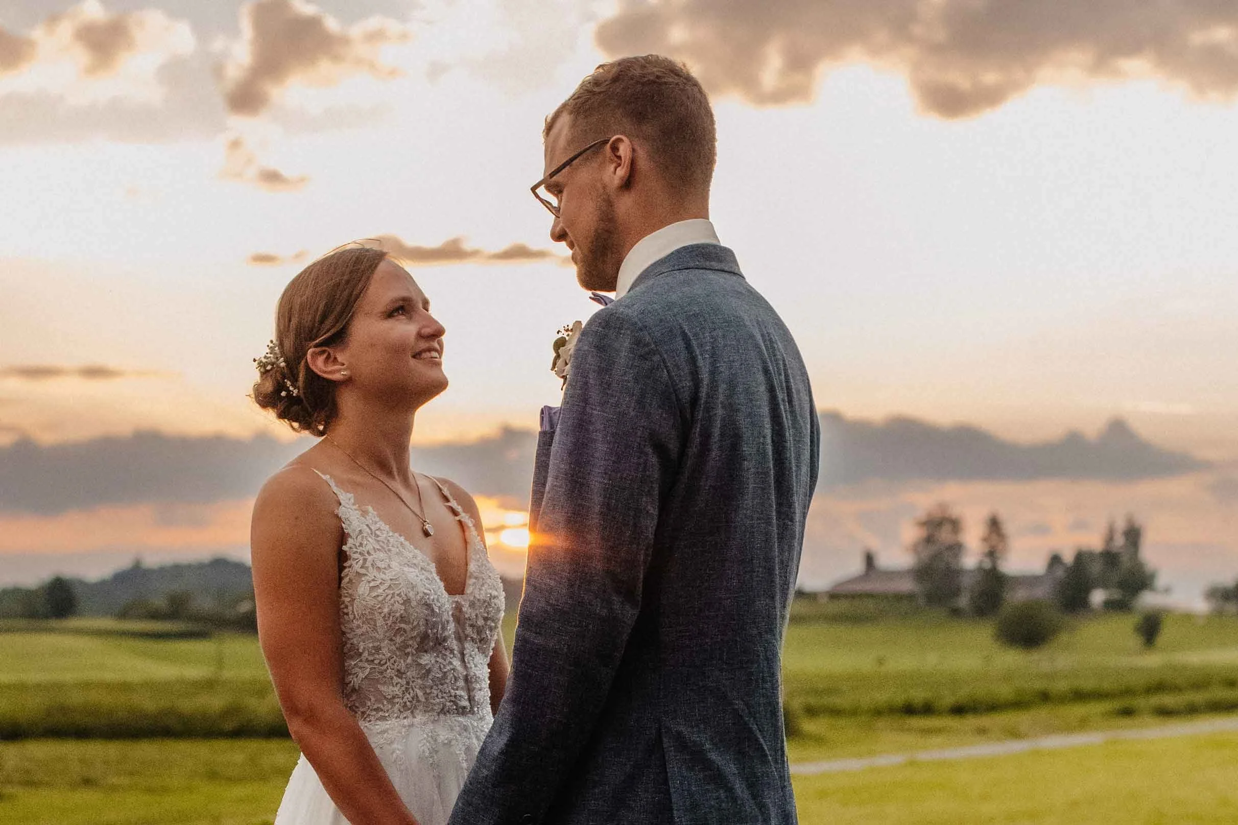 Brautpaar steht sich bei Sonnenuntergang auf einer grünen Wiese gegenüber, sie lächelt ihn an, er trägt einen blauen Anzug, sie trägt ein weisses Hochzeitskleid mit Spitze.