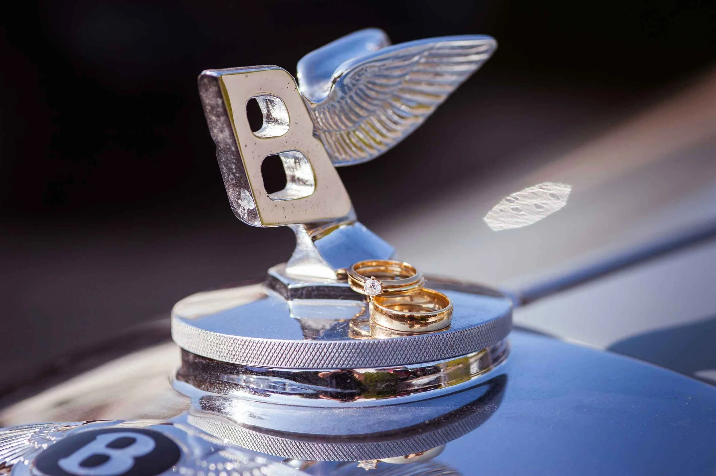 Zwei goldene Eheringe liegen auf einem silbernen Bentley-Emblem mit Flügel.