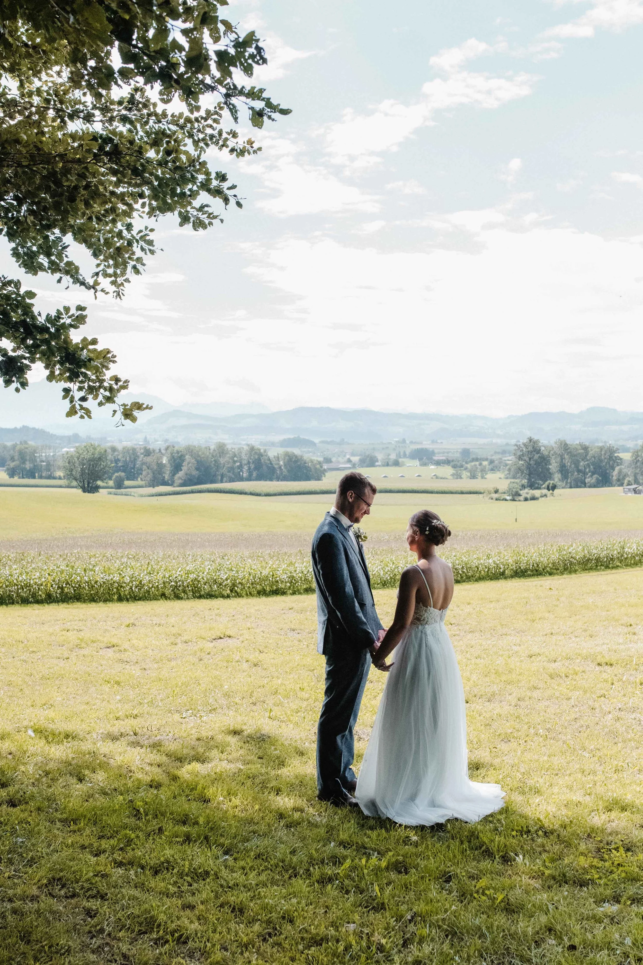 Brautpaar hält Händchen auf einer Wiese, im Hintergrund eine weite ländliche Landschaft mit Feldern und Bäumen, unter einem hellblauen Himmel mit wenigen Wolken.