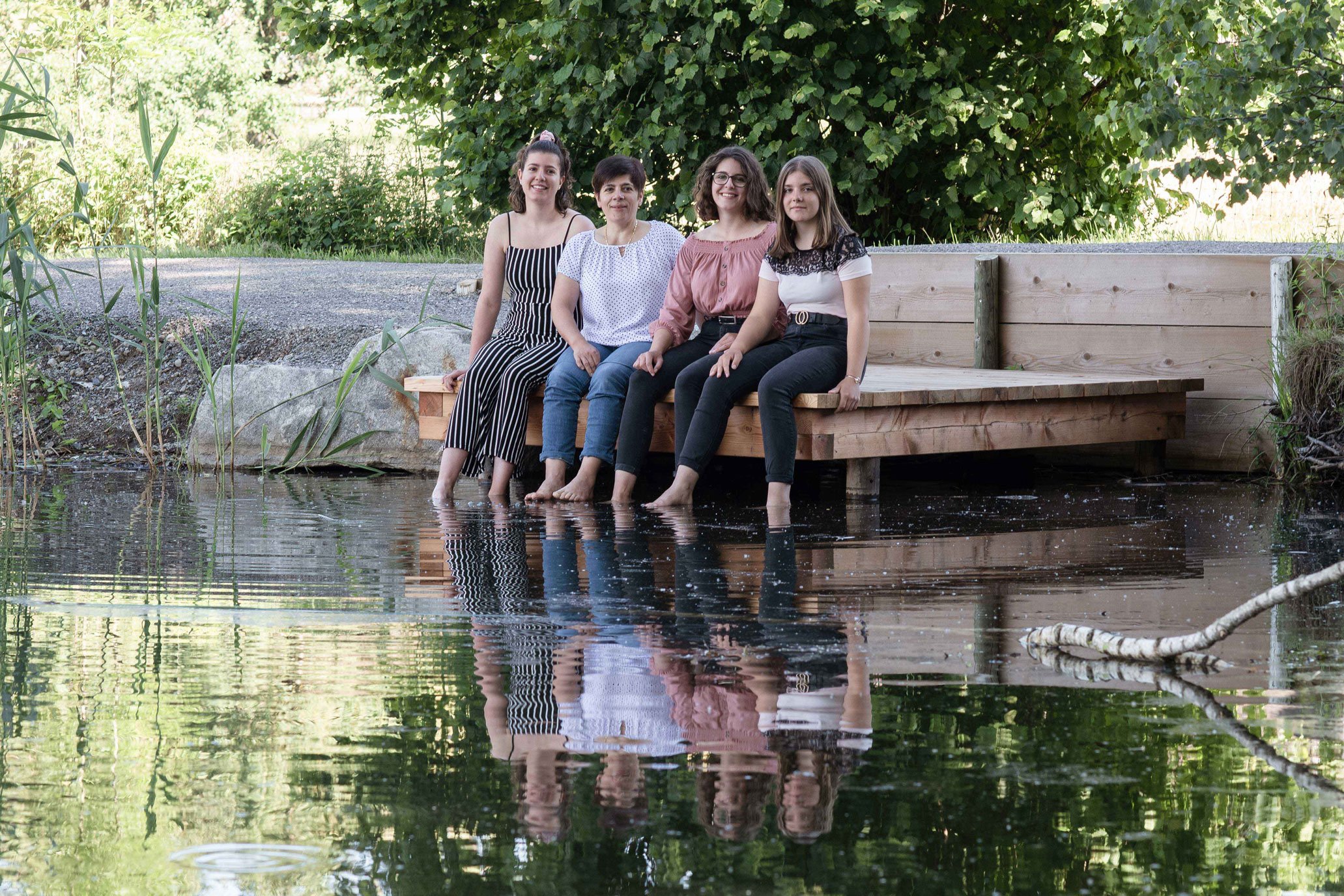 Mutter mit ihren Töchtern sitzen auf einem Holzsteg über einem Teich, ihre Füsse im Wasser, umgeben von grüner Vegetation.