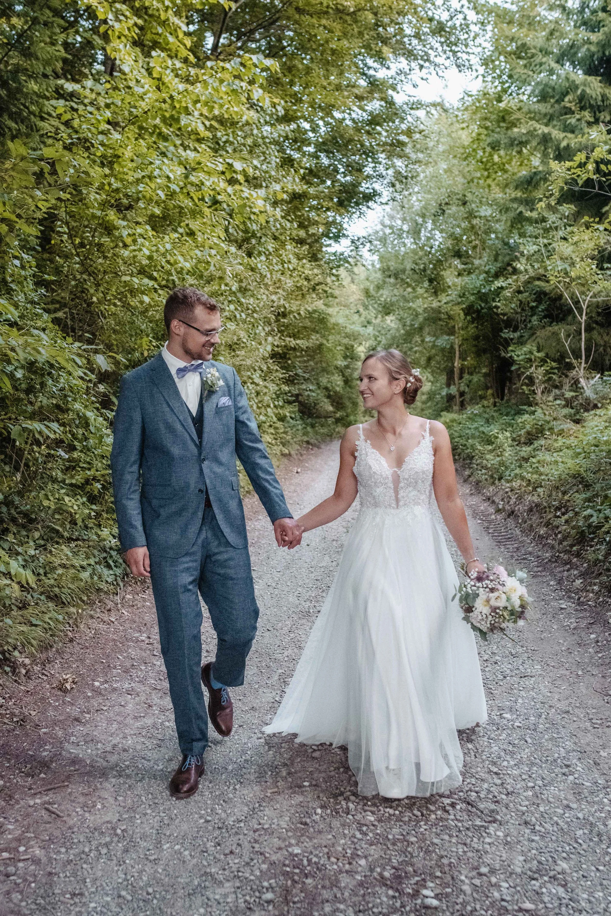 Brautpaar in Hochzeitskleidung läuft Hand in Hand durch einen Waldweg.