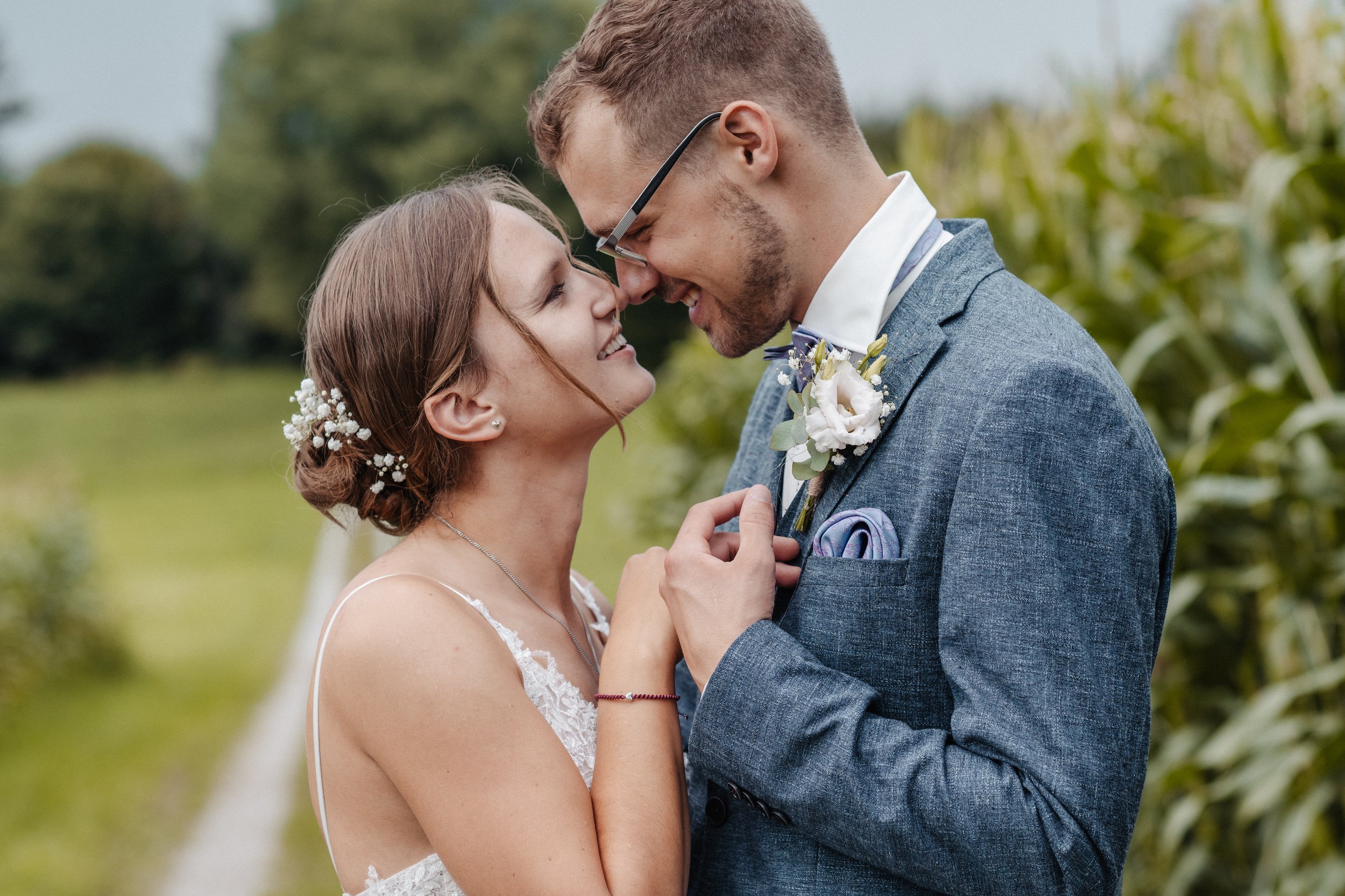 Hochzeitspaar steht einander zugewandt, lächelnd im Freien. Die Braut trägt ein weisses Kleid mit Spitzen und eine Blumenfrisur. Der Bräutigam trägt einen blauen Anzug mit Ansteckblume.