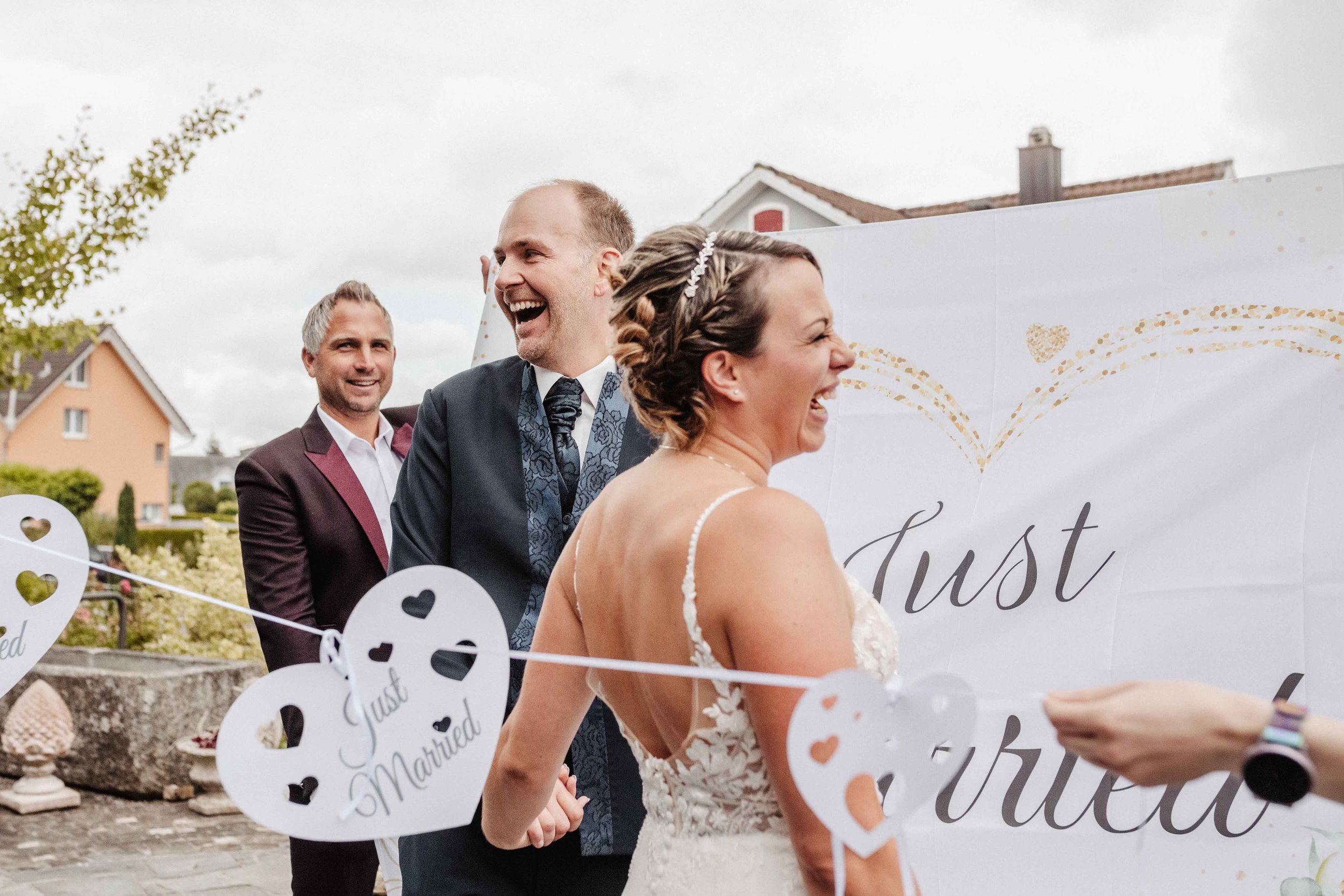 Hochzeitspaar lacht herzhaft. Girlande mit Herzen und 'Just Married'-Schilder im Vordergrund.