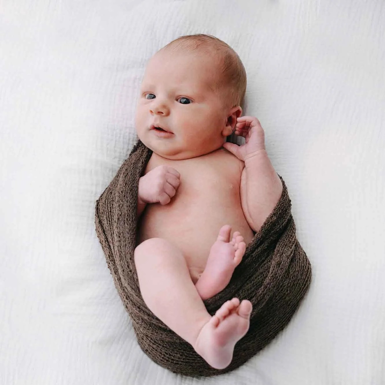 Ein neugeborenes Baby liegt auf einem weissen Tuch, mit einem braunen Tuch um den Körper, und schaut neugierig zur Seite.