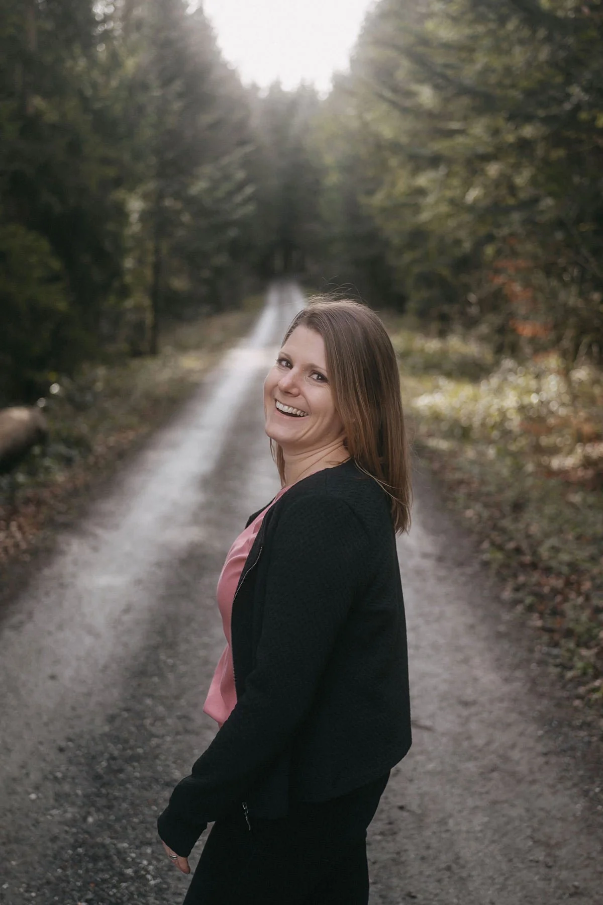 Junge Frau im Wald, lächelnd, trägt schwarze Jacke und pinkes Oberteil