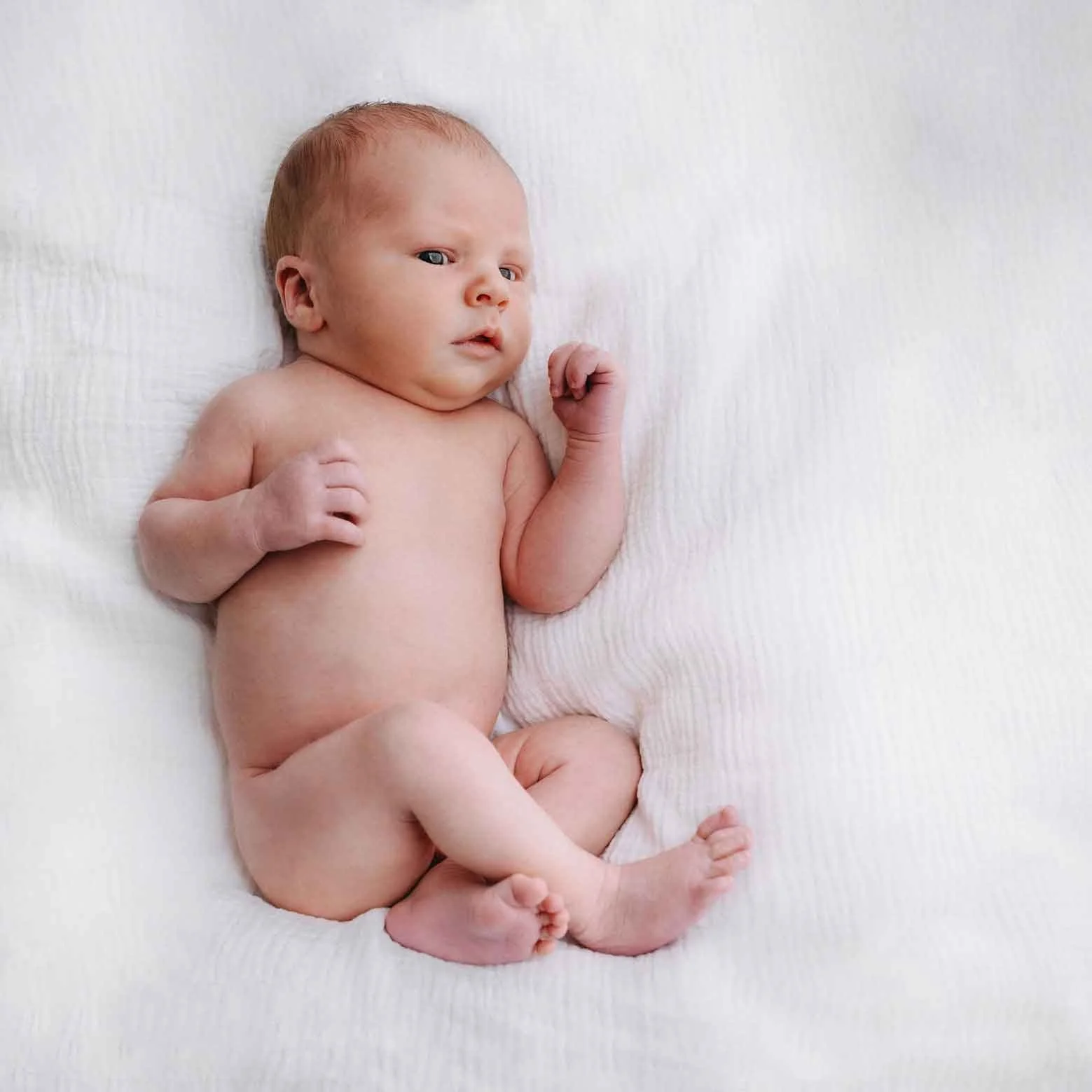 Baby liegt auf weissem Tuch, nackt, mit leichtem Blick nach links.
