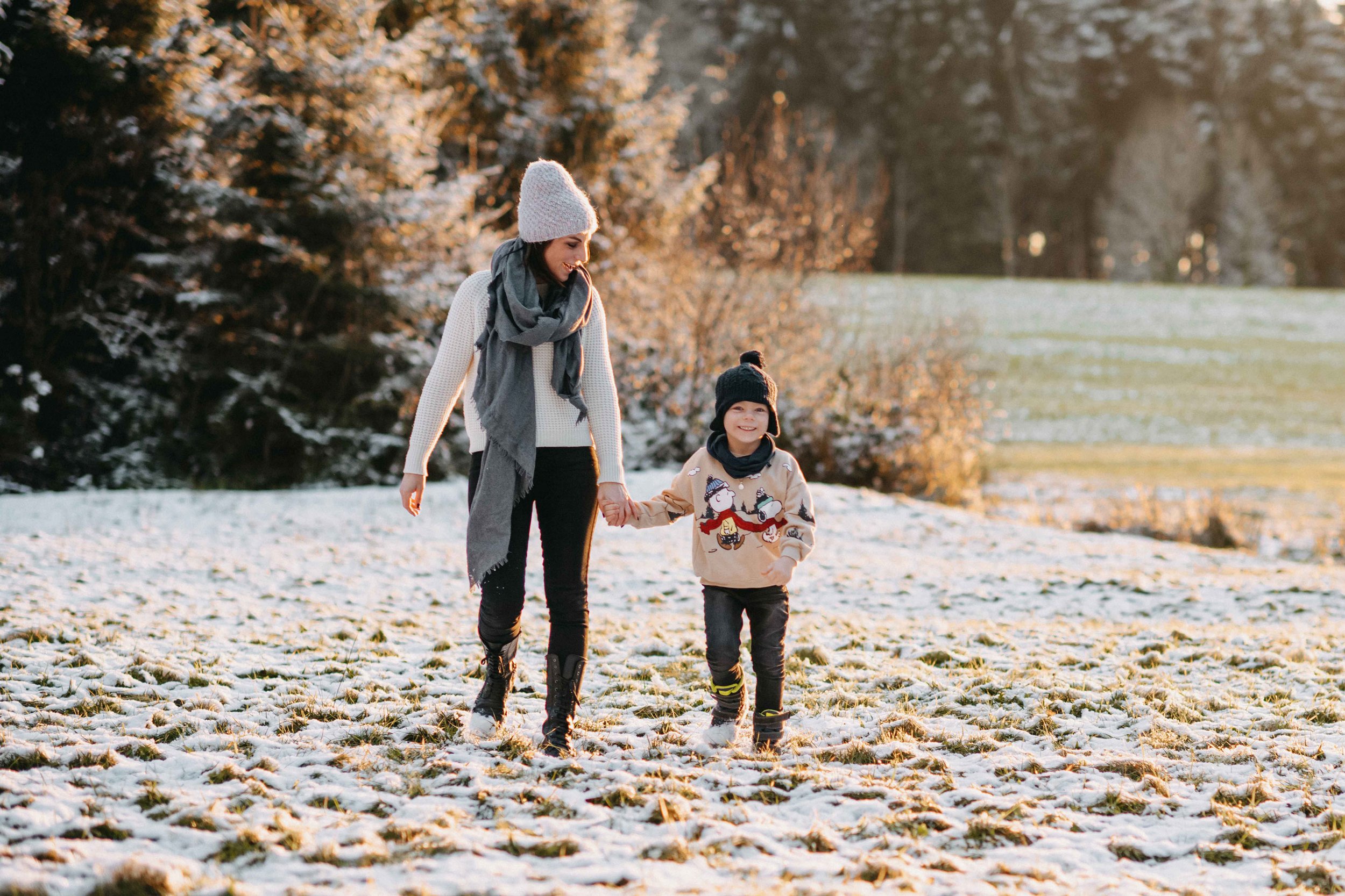 Mama und Kind spazieren im Schnee auf einem Feld, tragen Winterkleidung und lachen, mit Bäumen im Hintergrund. Sunset-Stimmung
