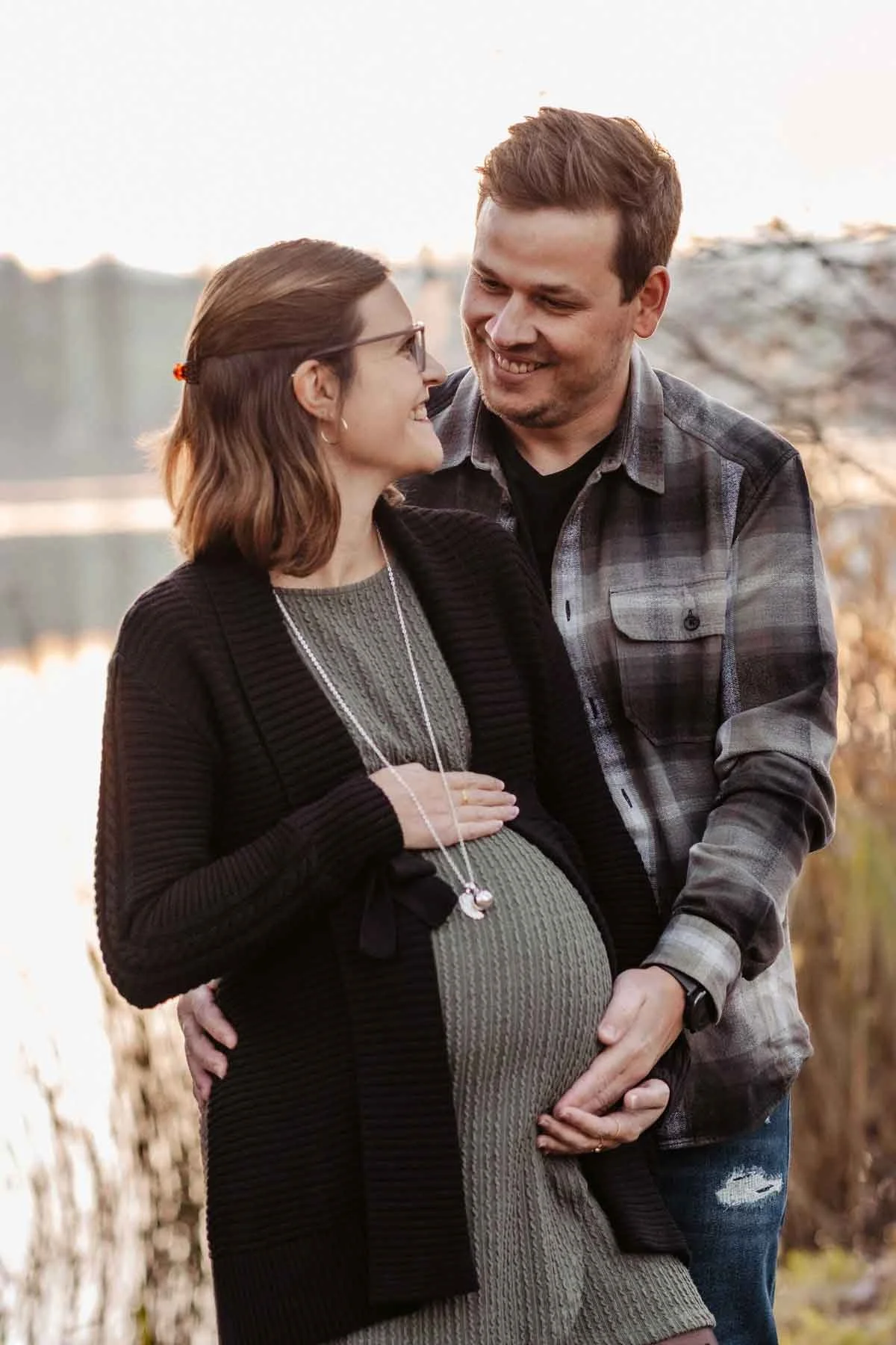 Ein schwangeres Paar steht am Wasser, lächelt sich an.