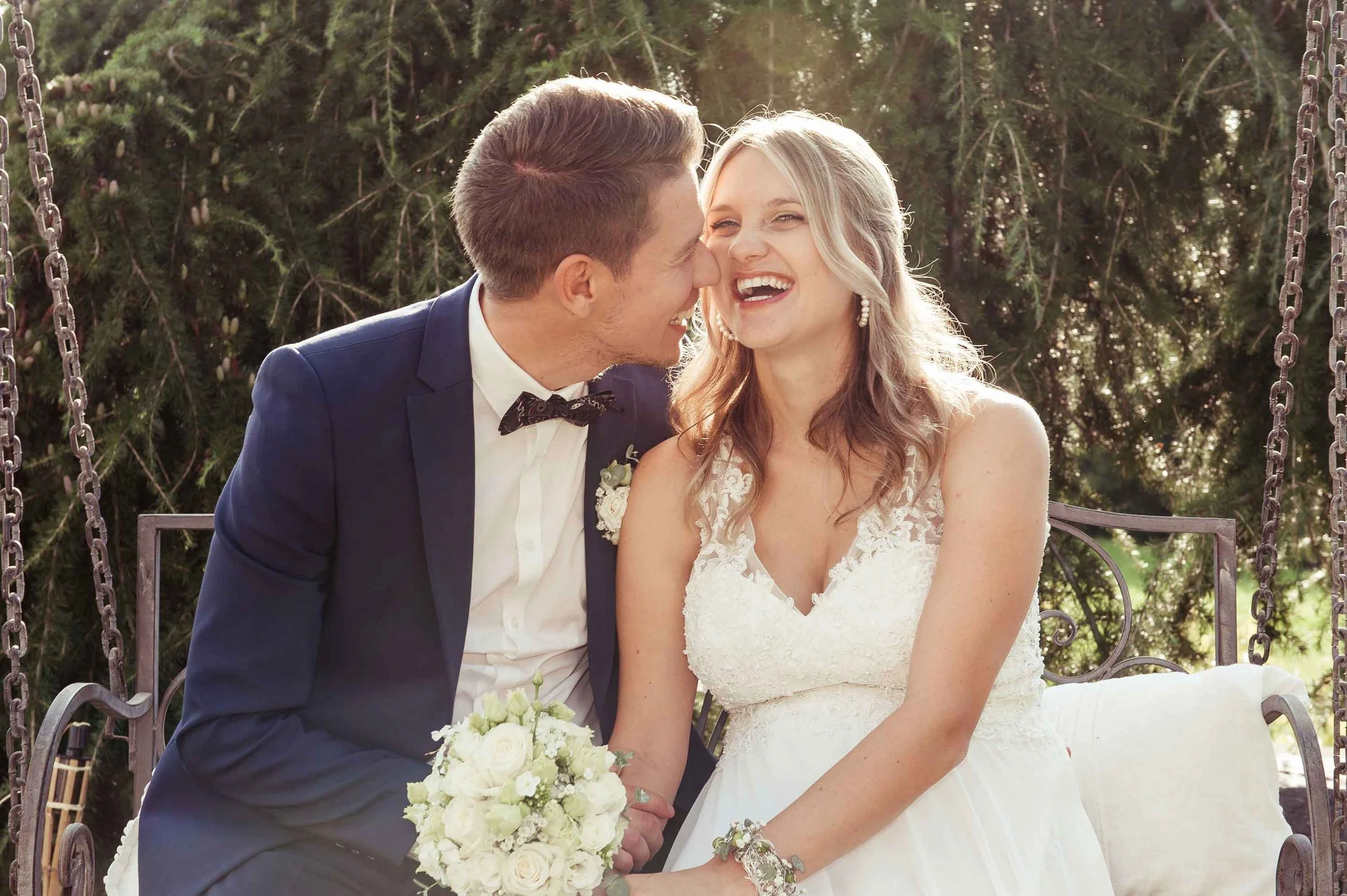 Ein glückliches frisch verheiratetes Paar sitzt auf einer Schaukel im Garten, umgeben von grünen Bäumen, lacht und geniesst den Moment.