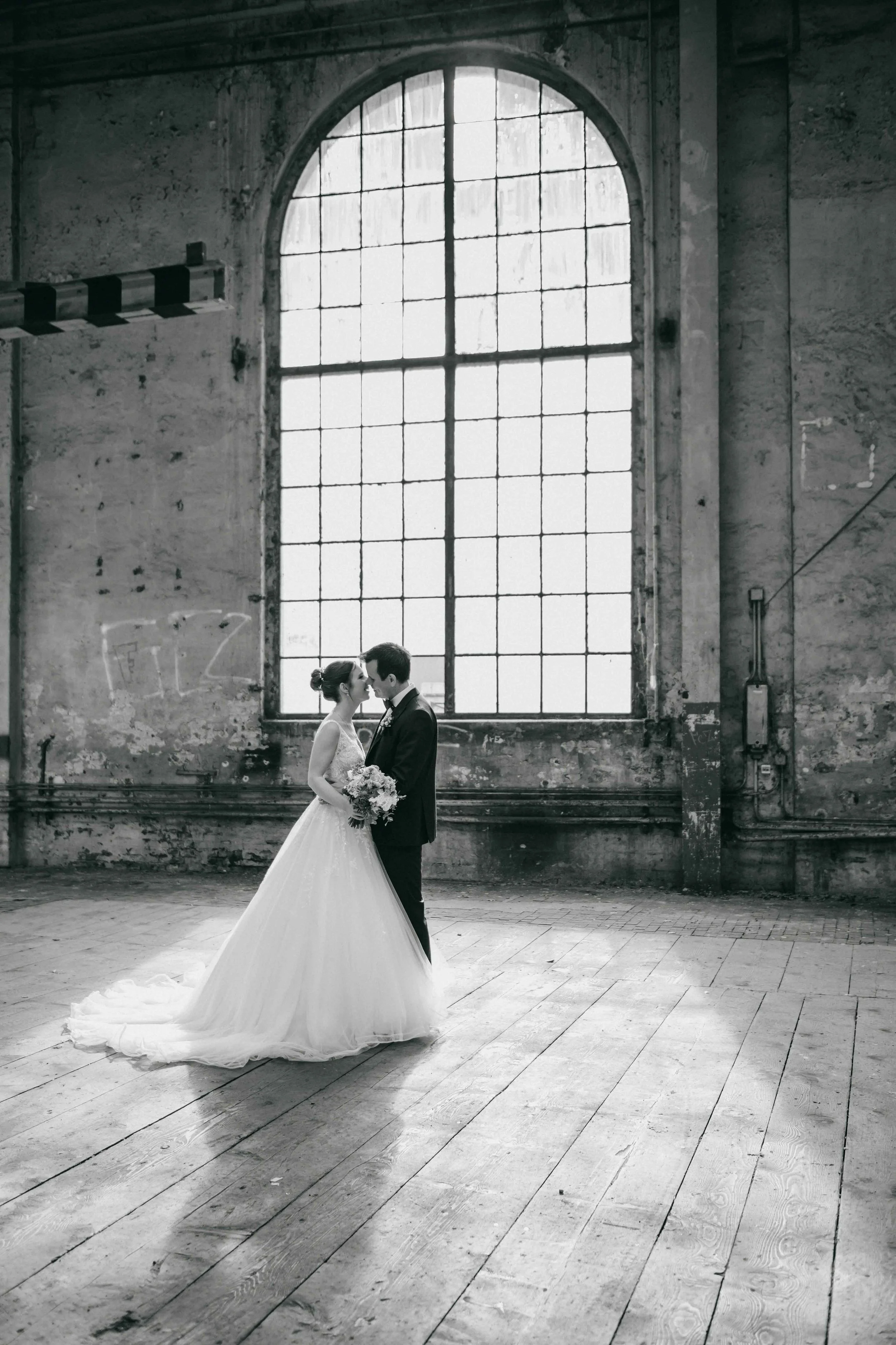 Ein Brautpaar in Hochzeitskleidung steht einander in einer alten, industriellen Räumlichkeit mit grossen Fenstern gegenüber und küsst sich.