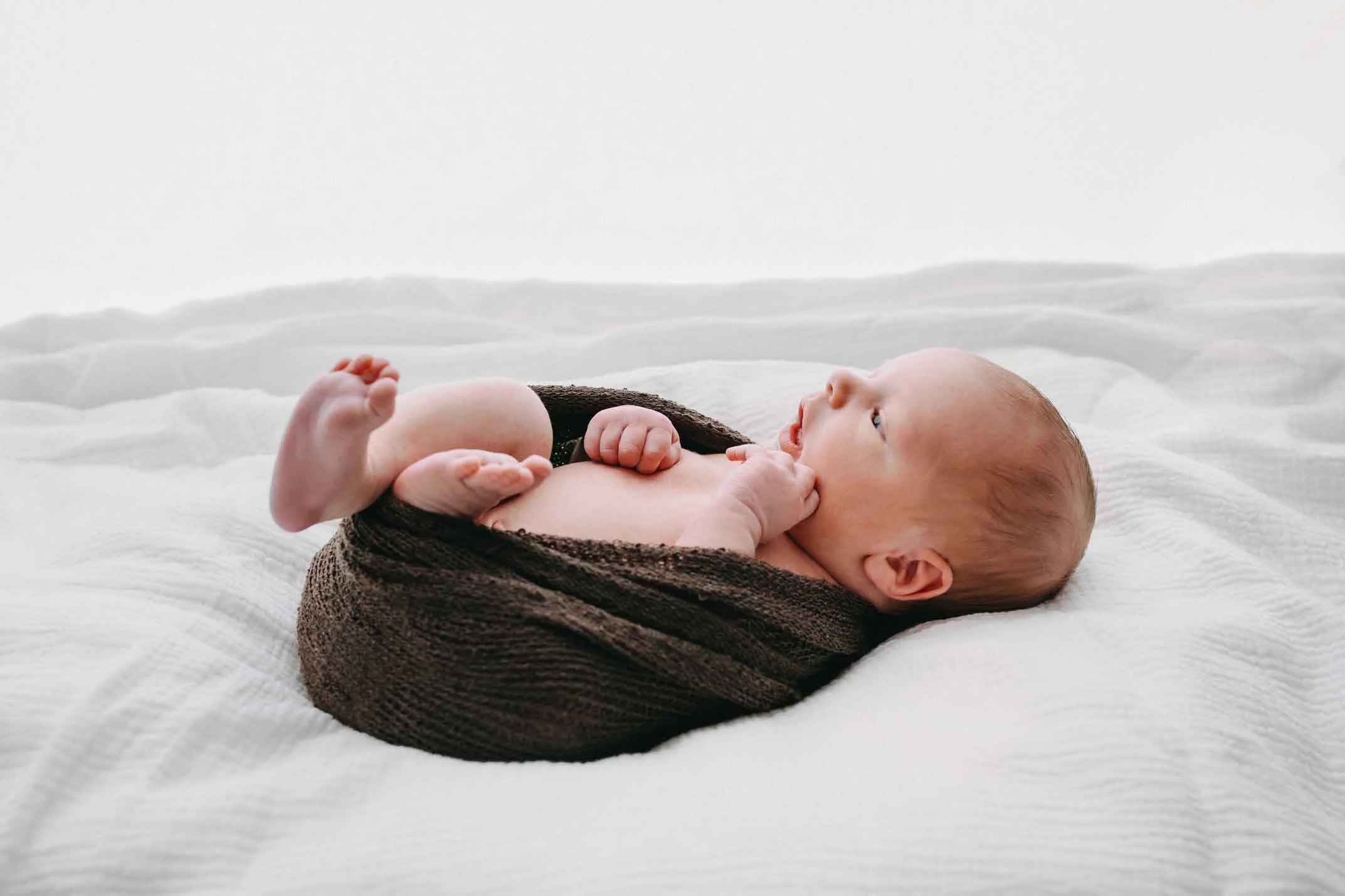 Neugeborenes Baby liegt auf einem weissen Bett, halb in eine braune Decke eingewickelt, und schaut nach oben.