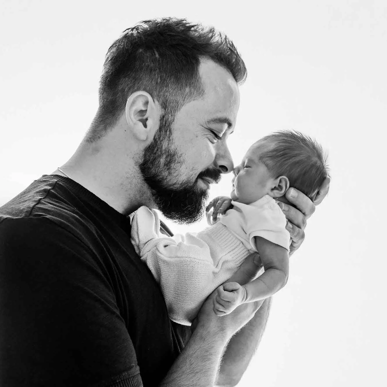 Ein Mann hält ein Baby liebevoll nahe an sein Gesicht, beide lächeln mit geschlossenen Augen.