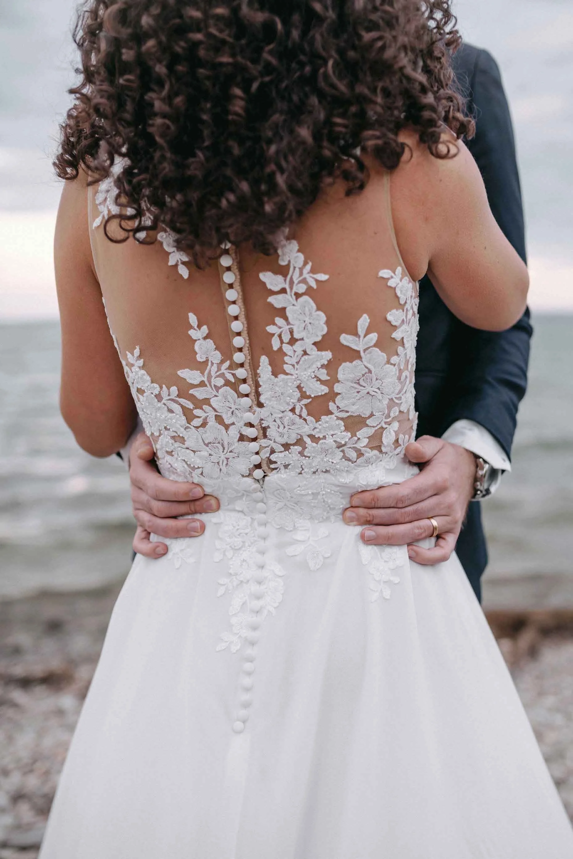 Nahaufnahme eines Hochzeitskleides mit floraler Spitze auf dem Rücken, die von einem Paar am Strand getragen wird.