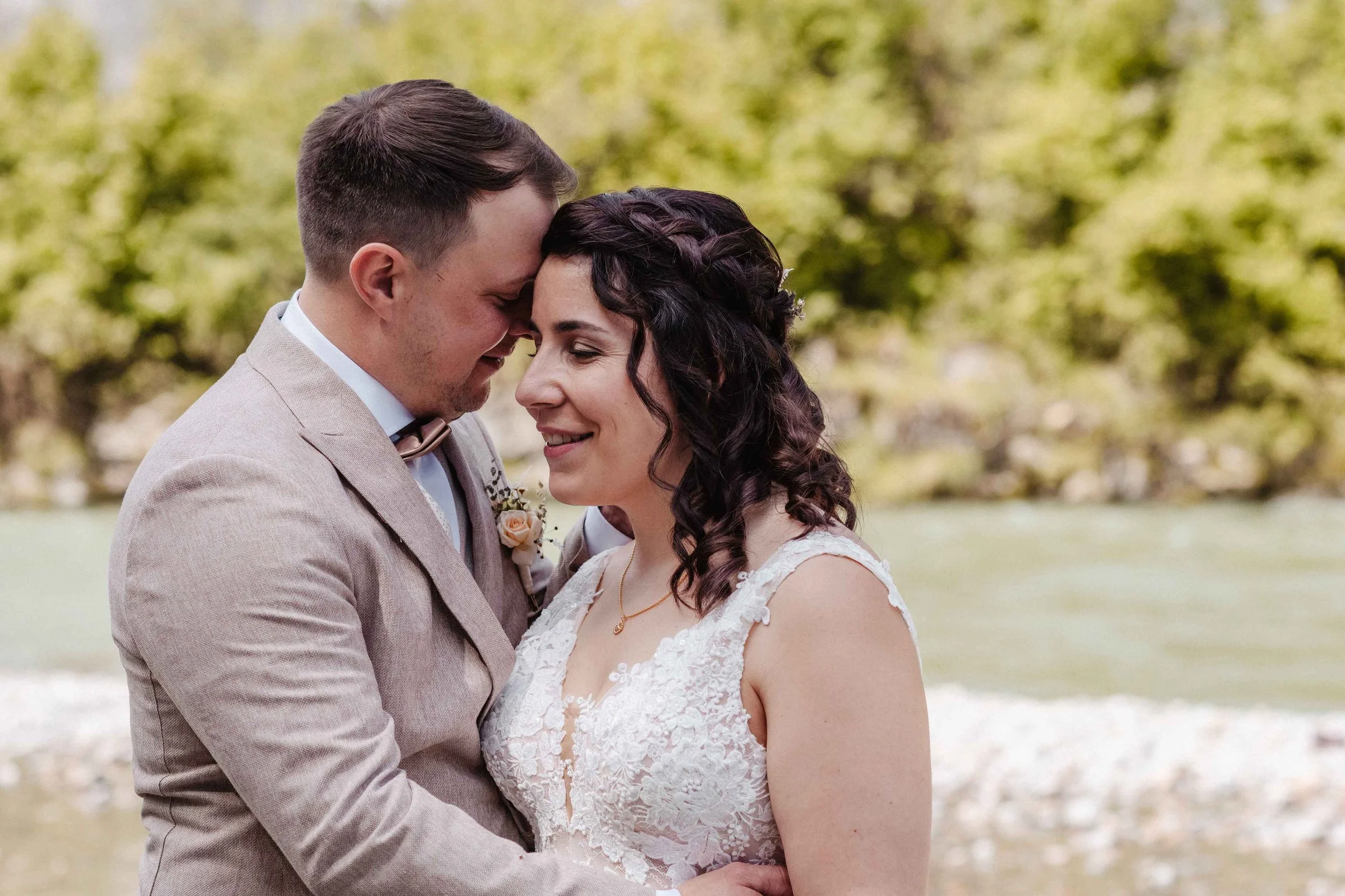 Brautpaar umarmt sich an einem Fluss, draussen in der Natur, an einem Tag mit sonnigem Himmel.