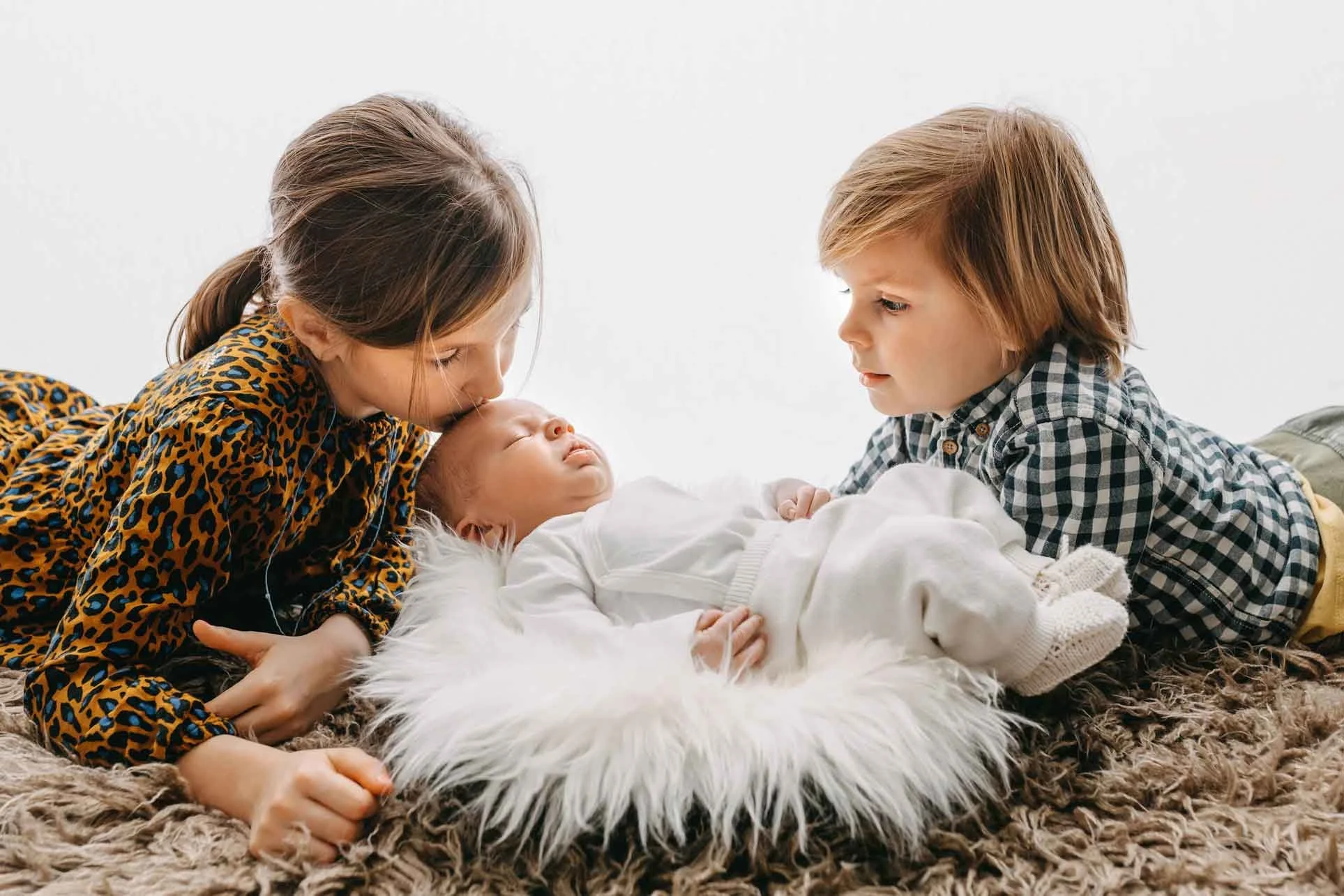 Zwei Kinder liegen auf dem Boden und betrachten ein Baby, das auf einem Kissen schläft. Ein Kind küsst das Baby auf die Stirn, das andere beobachtet es aufmerksam.