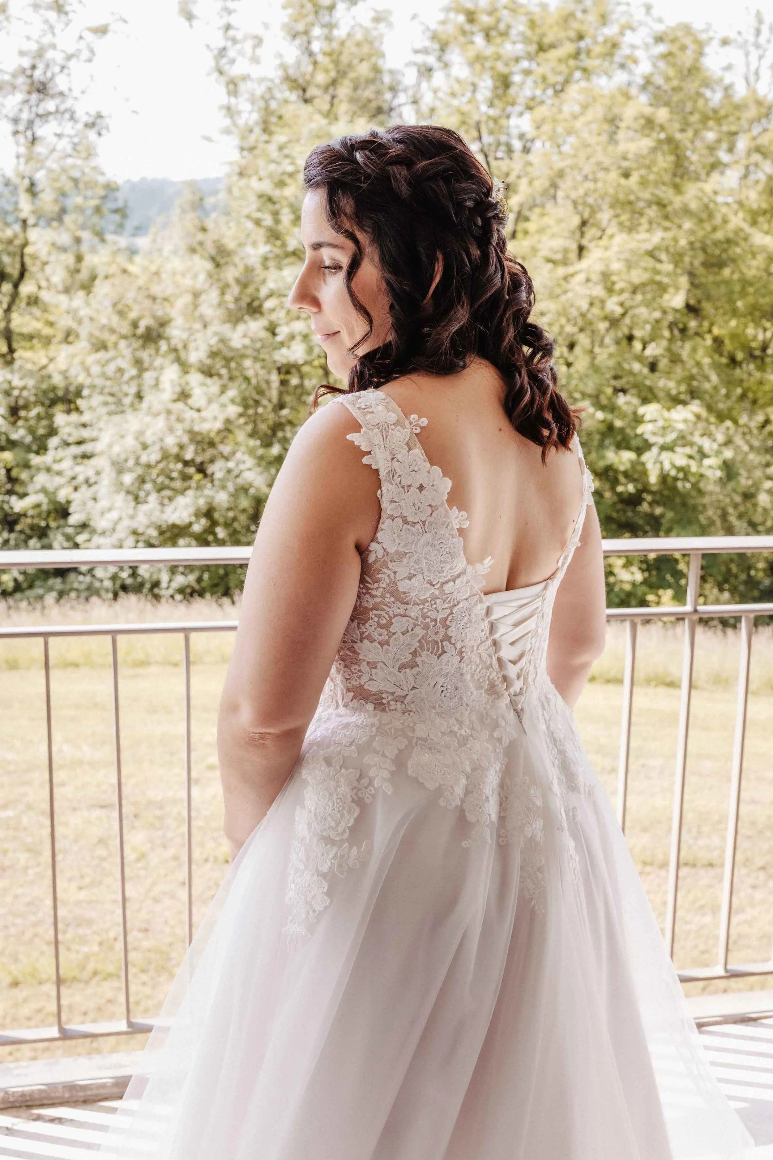 Braut in Brautkleid mit offenen Rücken auf Balkon mit grüner Landschaft im Hintergrund.