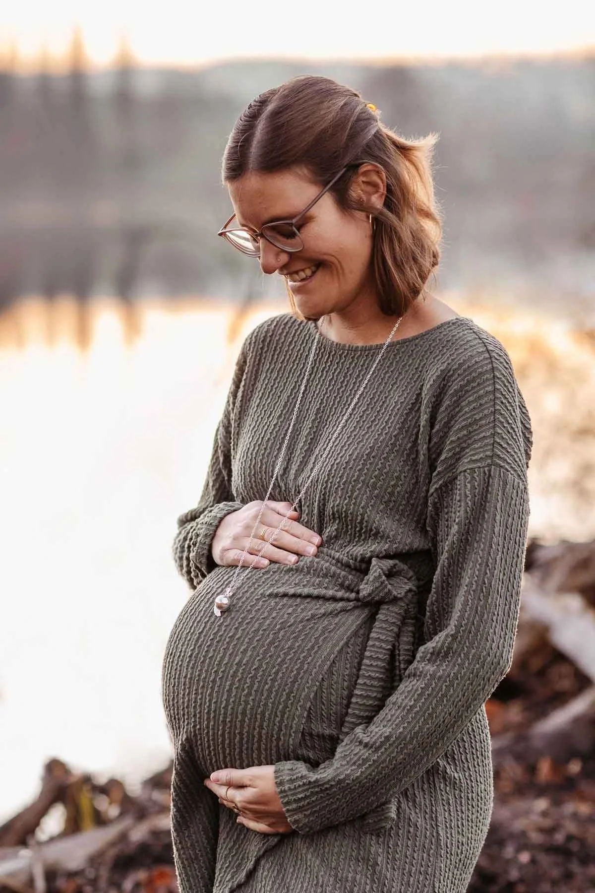 Schwangere Frau am Weiher bei Sonnenuntergang, hält den Bauch und lächelt.