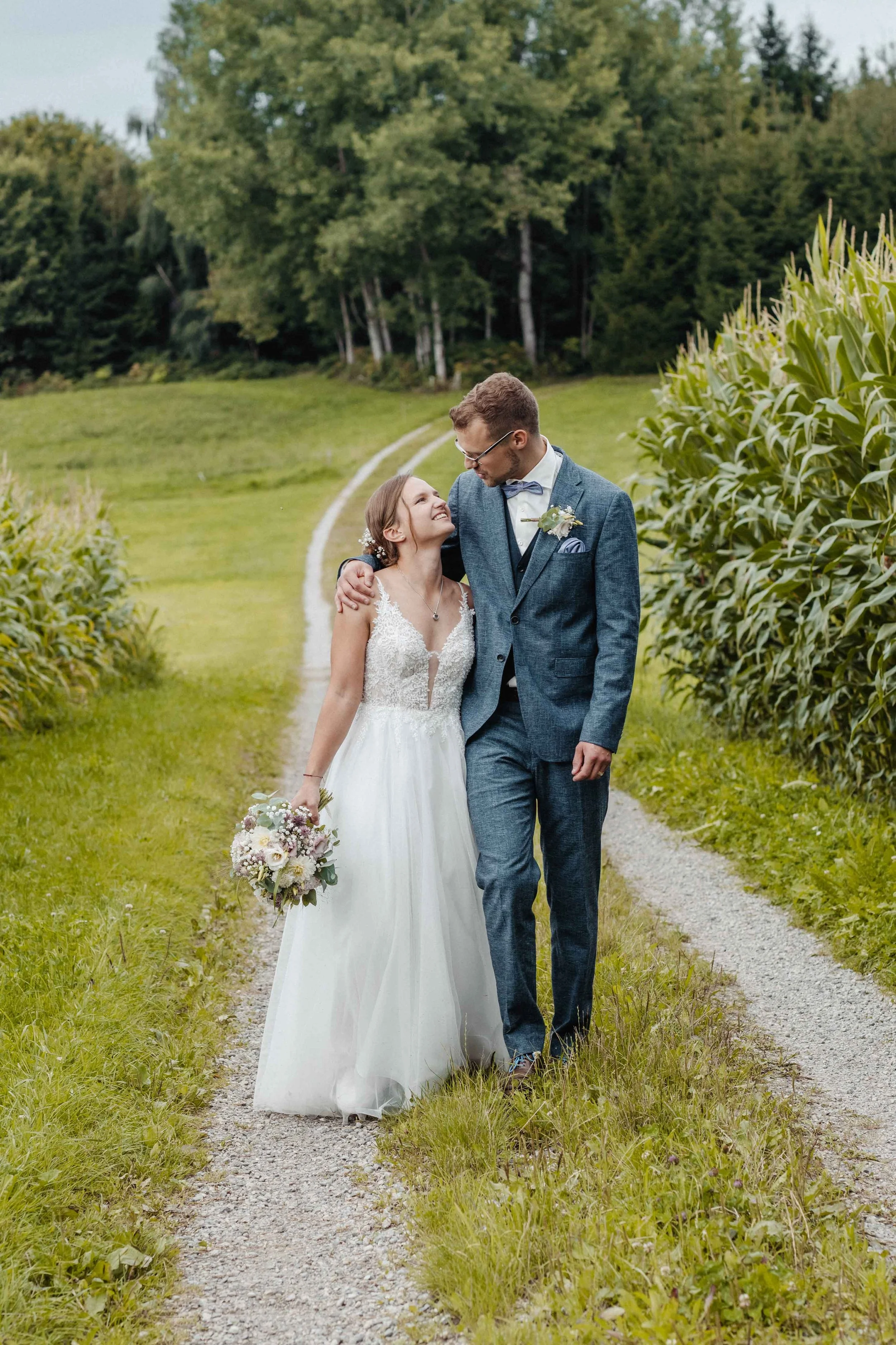 Braut in einem weissen Hochzeitskleid mit Brautstrauss, der Bräutigam in einem blauen Anzug mit Krawatte, gehen Hand in Hand entlang eines landwirtschaftlichen Weges in einer grünen, ländlichen Umgebung mit Bäumen und Maisfeldern.