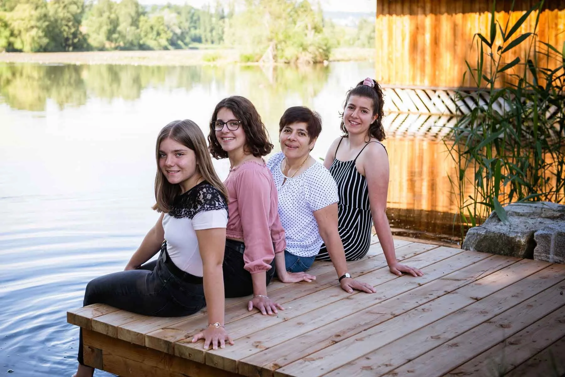 Mutter mit Töchter sitzen an einem Steg am See, lächeln und schauen in die Kamera.