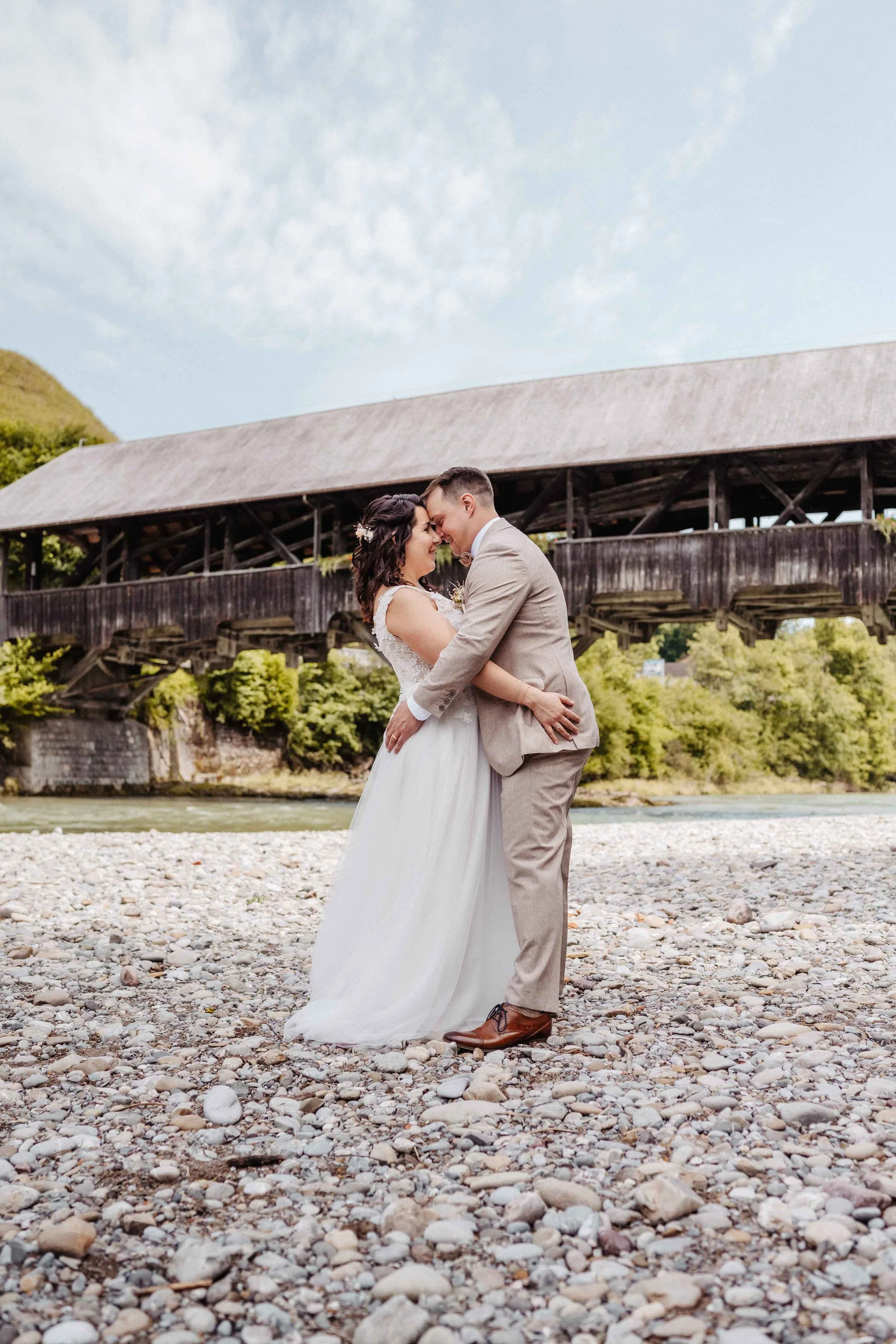Brautpaar, das sich herzlich umarmt, vor einer alten Holzbrücke bei schönem Wetter.