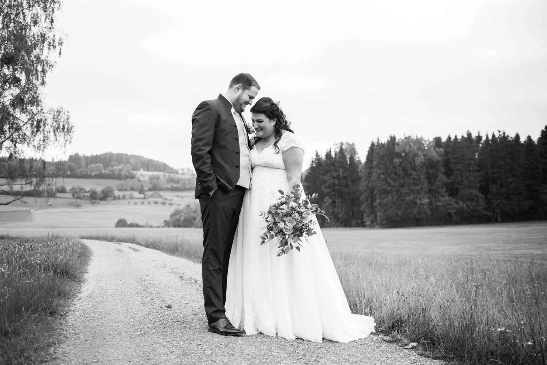 Braut und Bräutigam stehen sich auf einem Weg in einer ländlichen Umgebung gegenüber, die Braut hält einen Blumenstrauss, alles ist in Schwarzweiss.