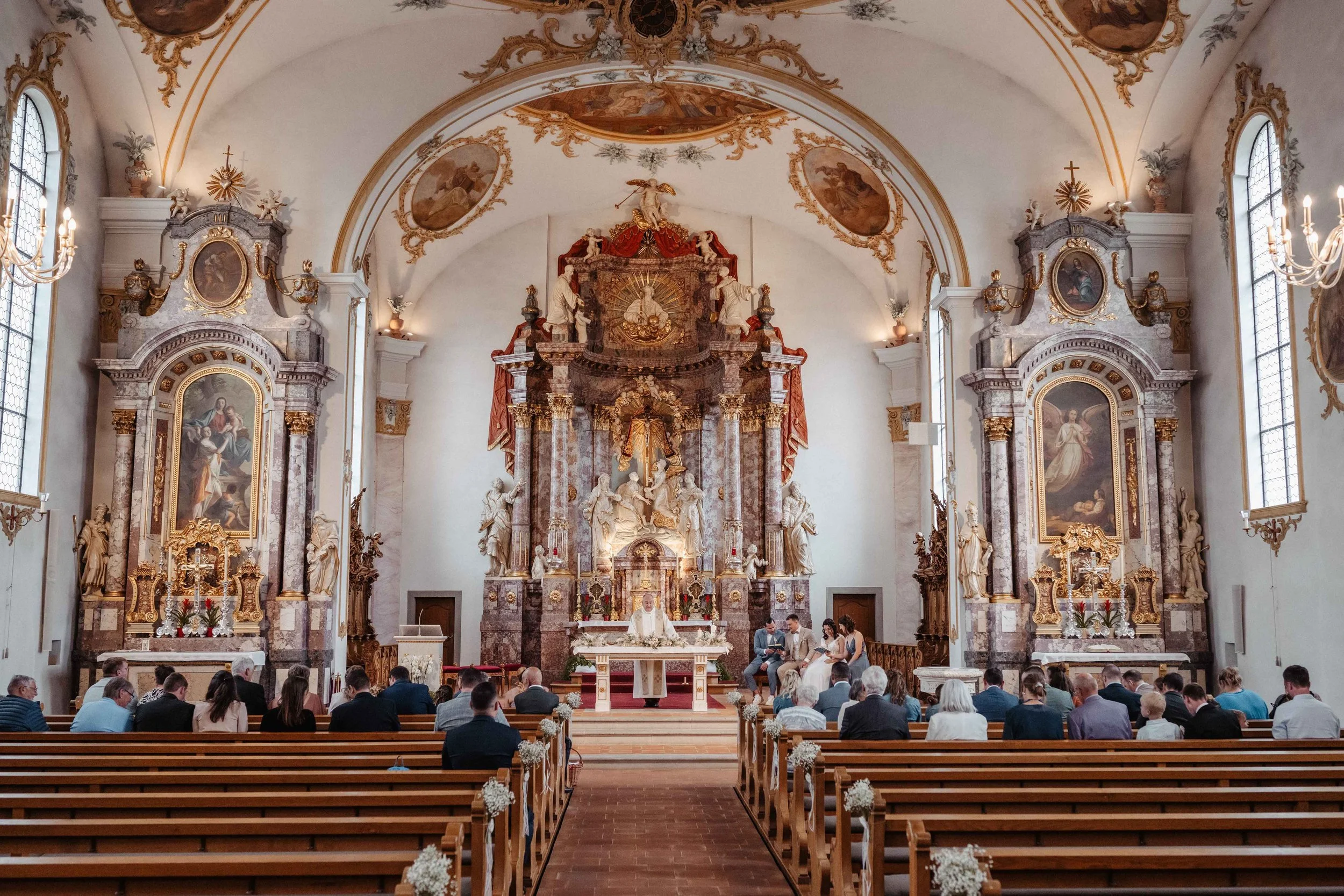 Innenraum einer Kirche bei der Hochzeitszeremonie mit Menschen auf Bänken, dem Altar und barocken Verzierungen.