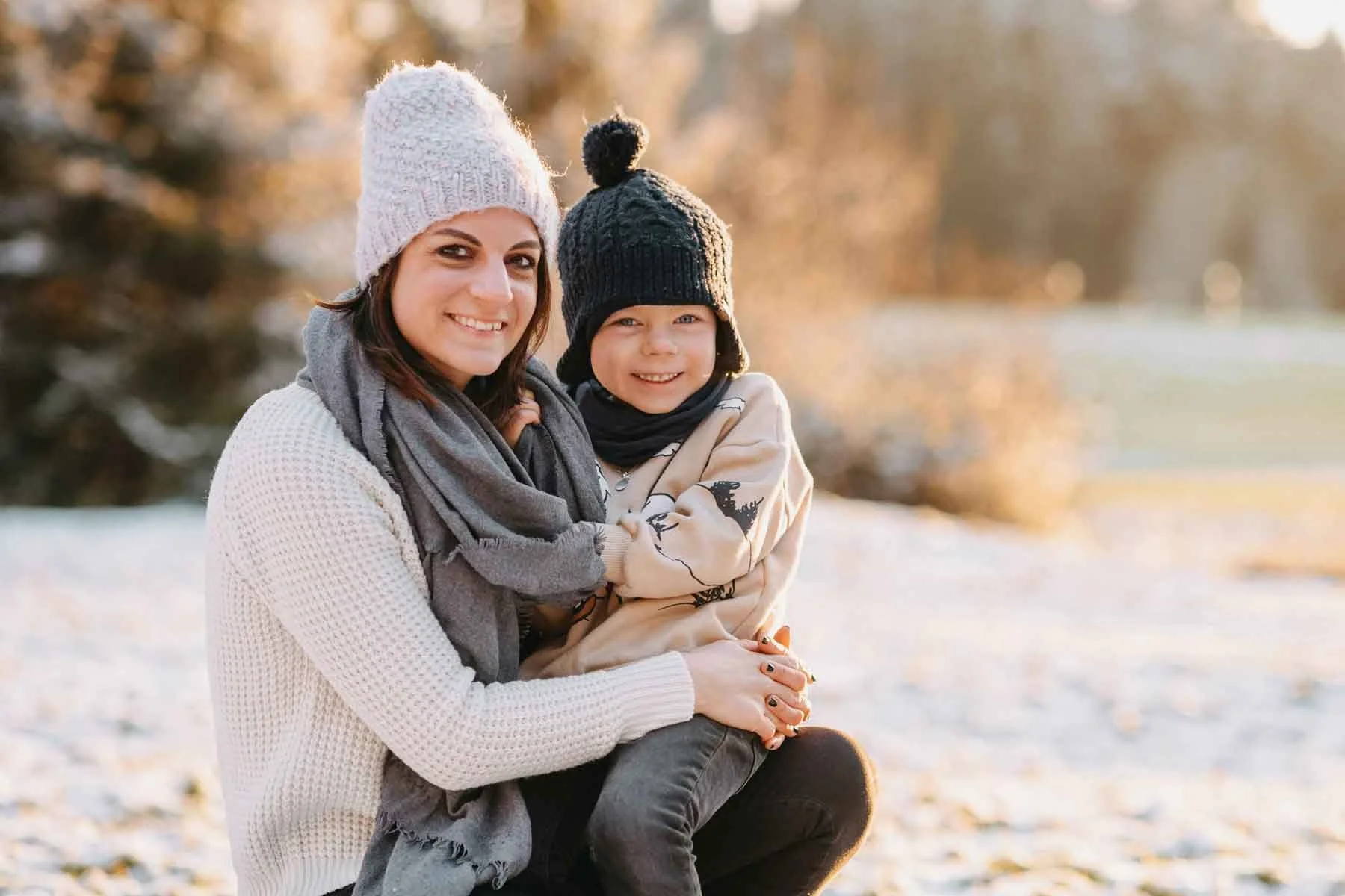 Mama mit Kind im Winter draussen, beide tragen Mützen und warme Kleidung, die Sonne scheint im Hintergrund.