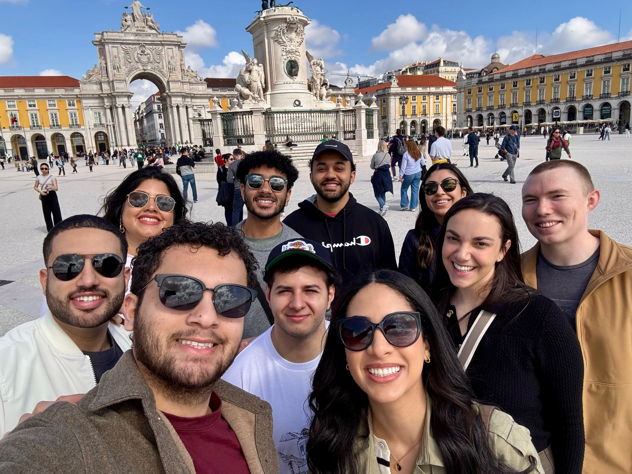 A group of ten diverse friends taking a selfie in a historic European square with yellow and white buildings, a large fountain, and a crowd of tourists.